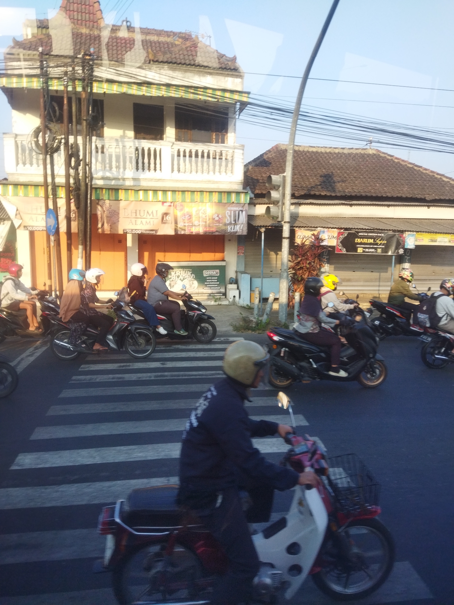 The image captures a street scene in Singodutan, Indonesia, with a focus on pedestrian crossings and vehicular traffic. In the foreground, a person wearing a dark jacket and a light brown helmet is riding a white and red scooter. The scooter has a basket on the front. The scooter is on a pedestrian crossing, indicated by white stripes on the asphalt. Behind this scooter, another scooter is partially visible, with a person wearing a black helmet and a dark jacket riding it.
In the mid-ground, several people are on motorcycles. From left to right, a person in a white jacket and red helmet is on a motorcycle. Next, a person wearing a brown hijab and a blue helmet is on a motorcycle, followed by a person in a white helmet and dark clothing. Further to the right, a person in a gray shirt and dark pants is riding a motorcycle. Behind them, another motorcycle with a rider in a yellow helmet is visible, and further back, a person in a green jacket and helmet is on a motorcycle.
The background shows buildings lining the street. On the left, there is a multi-story building with a white railing on its upper floor and a tiled roof. Below the railing, there are awnings with green and yellow stripes and signs with Indonesian text. On the right side of the street, a long, low-rise building with a brown tiled roof and open storefronts is visible. A traffic light is mounted on a pole in the middle of the scene. Overhead, a complex network of electrical wires is present. The sky is clear and bright, suggesting daytime. The overall scene depicts a busy urban environment with typical street activity in Indonesia.
