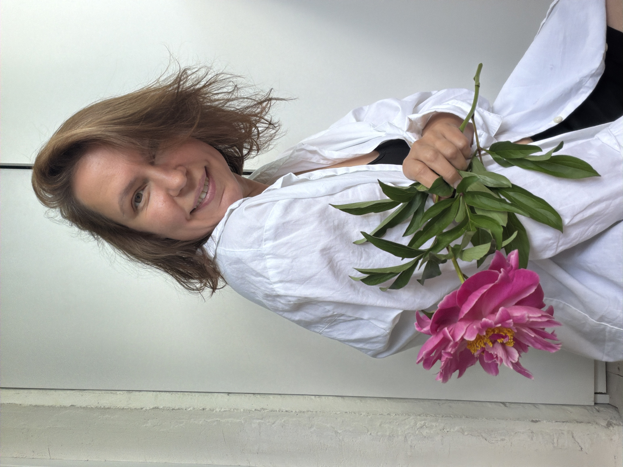A woman with medium-length brown hair is positioned on the left side of the frame, looking towards the camera with a slight smile. She is wearing a white button-up shirt, partially unbuttoned, revealing a black bra underneath. In her right hand, she holds a pink peony flower with green leaves. The background is a plain, light-colored wall with a vertical textured column on the far left, suggesting an indoor setting, possibly a home. The overall lighting is soft and natural. The location is not specified as Chaikovsky, Russia, or any other city.