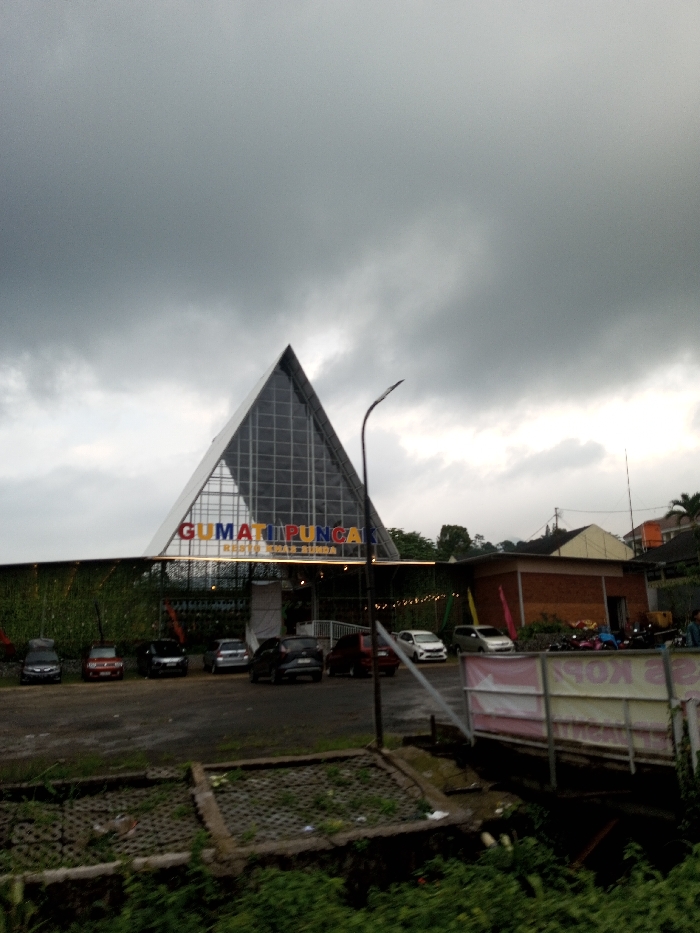 This image, identified by the user as located in Dawathang_Dorjibi_ Kashingtsawa, Bhutan, depicts an outdoor scene featuring a commercial establishment under an overcast sky.
The central subject is a large building with a distinctive triangular, possibly pyramidal, glass facade forming its upper structure, characterized by a visible grid pattern. Below this glass section, a horizontal sign displays the multi-colored text "GUMATI PUNCAK" in prominent letters, with "RESTO KHAS SUNDA" written directly beneath it. The lower section of the building features a wall adorned with green foliage or a vertical garden, and a line of string lights is visible along its length. An entrance area with white architectural elements is positioned beneath the sign.
In front of the building, a dark-paved parking lot contains at least seven parked passenger vehicles of various models and colors, including sedans and a minivan. A tall, dark, curved street lamp post with a modern fixture stands in the mid-ground, slightly to the right of the building's center. To the right side of the main building, a smaller, single-story brown brick building with a reddish roof is visible, with multiple motorcycles parked in its vicinity. Further in the background, additional buildings and dense green trees are present.
The immediate foreground consists of uneven terrain covered with wild green vegetation and some concrete block structures. A partially obscured white banner with faint red text is visible on the far right. The sky is entirely overcast with heavy grey clouds, resulting in dim, diffused lighting across the entire scene. No individuals are clearly discernible, and no specific actions or interactions are depicted.