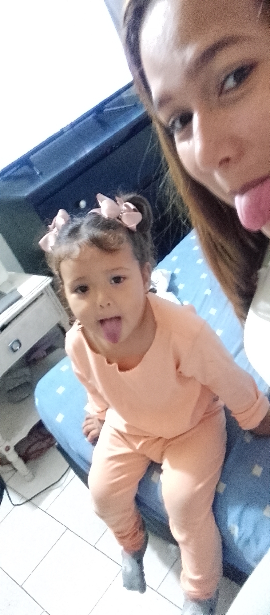 A woman and a young girl are posing for a selfie, sticking their tongues out. The woman is in the foreground, her face and part of her torso visible. She has long, brown hair and her tongue is extended. The young girl is seated in front of her, also sticking her tongue out. She has curly brown hair styled with two light pink bows and is wearing a peach-colored long-sleeved shirt and pants. A gray sock is visible on her right foot.
The setting appears to be indoors, likely a bedroom. A blue and white checkered surface, possibly a bedspread, is visible behind the girl. To the left of the girl, a white piece of furniture with a drawer and a metallic handle is partially visible, along with a white cable. In the background, a dark blue piece of furniture is present. The lighting suggests it is daytime. The overall mood is playful and lighthearted, with both subjects engaging in a fun gesture. The location context of Caracas, Venezuela is provided. No visible text is present in the image.