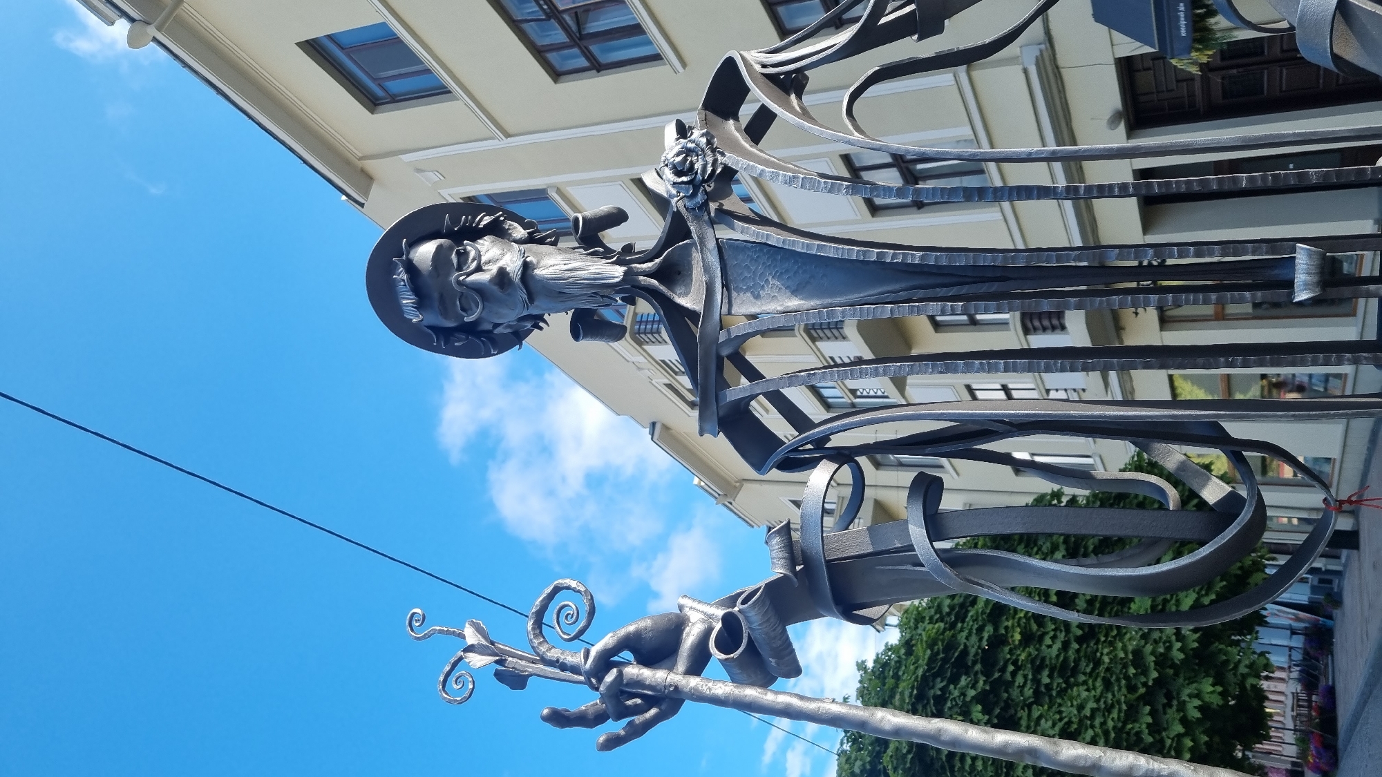 This is a close-up, low-angle shot of a metal sculpture of a man wearing a hat, glasses, and a beard, holding a staff. The sculpture is dark gray and appears to be made of wrought iron, with a detailed face and flowing beard. The man is wearing a hat with a brim, and has a pipe in his mouth. He is also holding a staff in his left hand, which has decorative spiral elements at the top. A metallic rose is attached to his chest.
In the background, a light-colored building with multiple windows and balconies is visible. The building has a modern architectural style. Above, a bright blue sky with a few white clouds is seen, with a thin black wire or cable stretching across the frame.
To the left of the sculpture, a lush green tree with dense foliage is partially visible, and further to the left, a street scene with flowerbeds and possibly shops can be observed.
This sculpture is located on Theater Square in Chernivtsi, Ukraine, and is a representation of the writer and poet Ivan Myronovych Melnychuk.