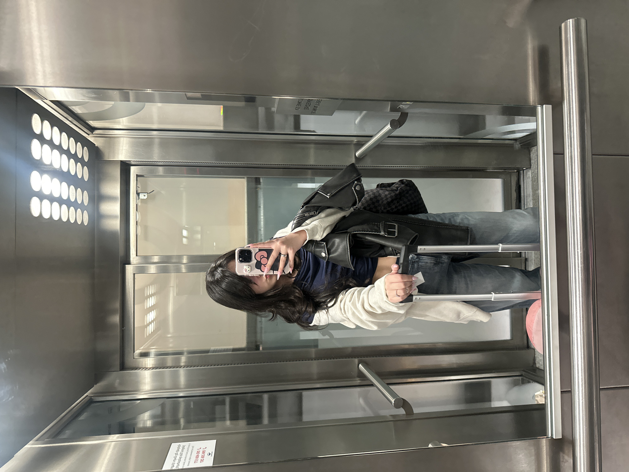 A young woman stands in an elevator, taking a selfie with her phone. She wears a black leather jacket over a navy blue top and jeans, along with a white sweater. A black patterned bag rests near her shoulder. The elevator has a modern, stainless steel interior, including round lights on one side. A luggage is with her. There is a sign in Armenian on the wall. The city location is not identifiable from the image.