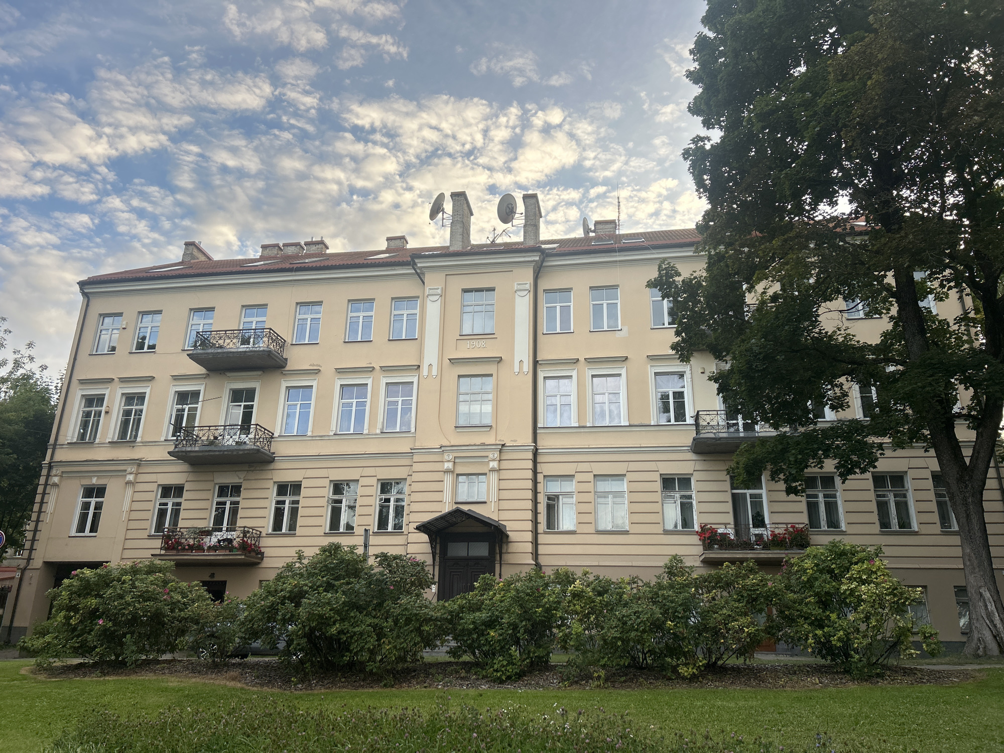 The image shows a building located at Antakalnio gatvė 11 in Vilnius, Lithuania. The building is a light yellow, multi-story apartment building with a red tiled roof. It has several windows on each floor, some with balconies. A large tree with dark green leaves is partially visible on the right side of the building. In front of the building, there is a lawn with some bushes and flowers. The sky is cloudy with patches of blue. The architectural style appears to be from the early 20th century. There are no people visible in the image.