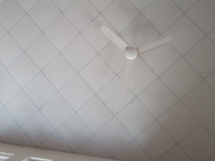 An interior view within the city of Ayama, Nigeria, depicts a white three-bladed ceiling fan centrally positioned in the upper-left quadrant. The fan is mounted on a grid of white square acoustic ceiling tiles, which display dark grout lines or a framing system. Several ceiling tiles exhibit minor dust accumulation or discoloration. A section of an interior wall, featuring multiple window panes, is visible along the right edge of the frame. No individuals or active subjects are present.