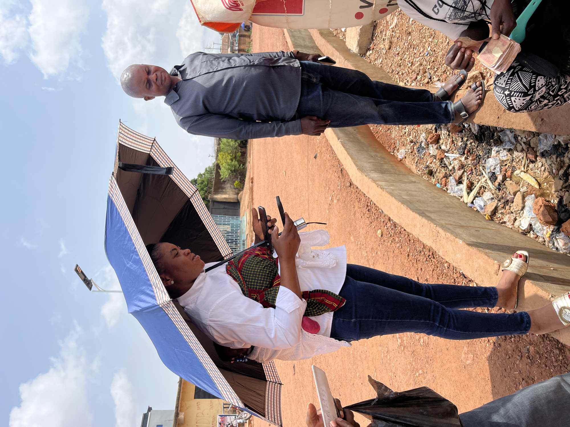 The image shows a woman sitting under a blue and black umbrella on a dirt ground in Gura Top, Nigeria. She wears a white shirt, jeans, and sandals, and holds a phone in her hands. A red and green fabric is draped across her body. Beside her are two men and garbage on the ground and one of the men is holding money. The background shows buildings and a bright sky. Another man standing is wearing a gray shirt and jeans.