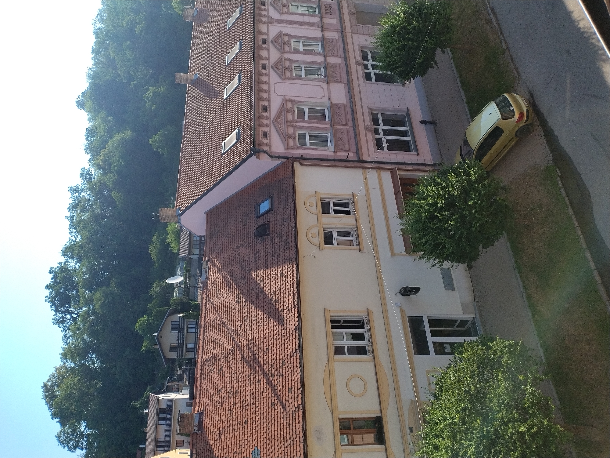 An aerial view captures a street scene in Hluboká nad Vltavou, Czechia, bathed in sunlight. Two buildings line the street; the one on the left is cream-colored with a terracotta tiled roof, featuring multiple windows and a storefront entrance. To its right, a pale pink building with intricate architectural details also has a terracotta tiled roof, punctuated by several skylights. A yellow hatchback car is parked on the cobblestone area beside the buildings. Lush green trees and manicured lawns flank the street and sidewalk. In the background, a dense forest covers a hill under a clear blue sky.