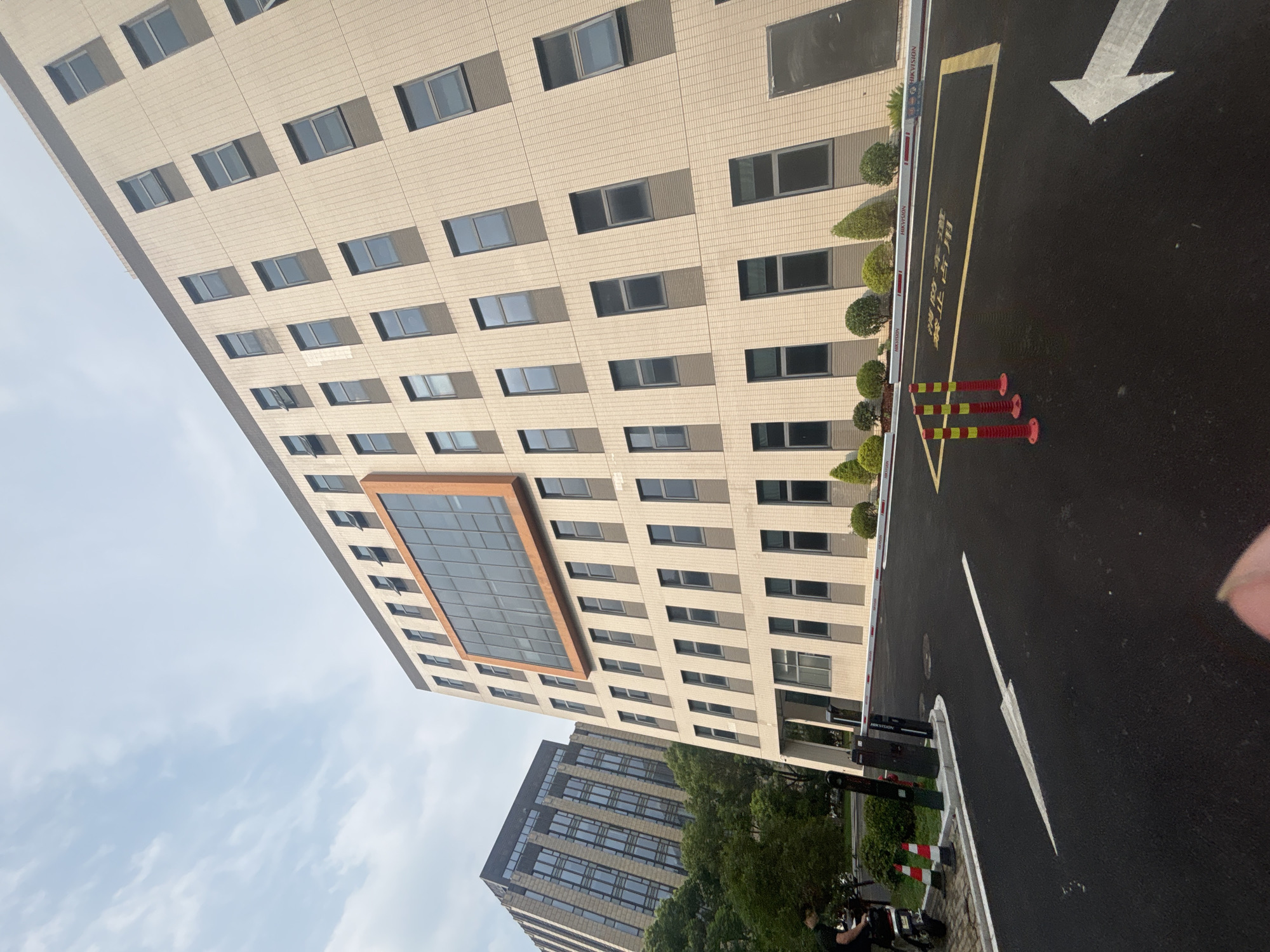 This is a ground-level, wide-angle photograph of the exterior of a modern office building located in Suzhou Industrial Park, China. The building is tall and appears to be constructed with beige tiled panels and dark gray window frames. A large, prominent orange-framed window or facade element is centered on the upper floors. Multiple rows of rectangular windows, some open, are visible across the building's facade. To the left of the main building, another taller office building with a glass and stone facade is partially visible, along with lush green trees. In the foreground, there is a paved asphalt area with white directional arrows painted on it, indicating traffic flow. Several red and yellow traffic bollards are positioned near a yellow painted rectangle on the asphalt, which is marked with Chinese characters. A white barrier gate is visible across the entrance, with a security camera and what appears to be a parking meter or access control system to its left. A person is seen on a scooter to the far left of the image. The sky is partly cloudy.