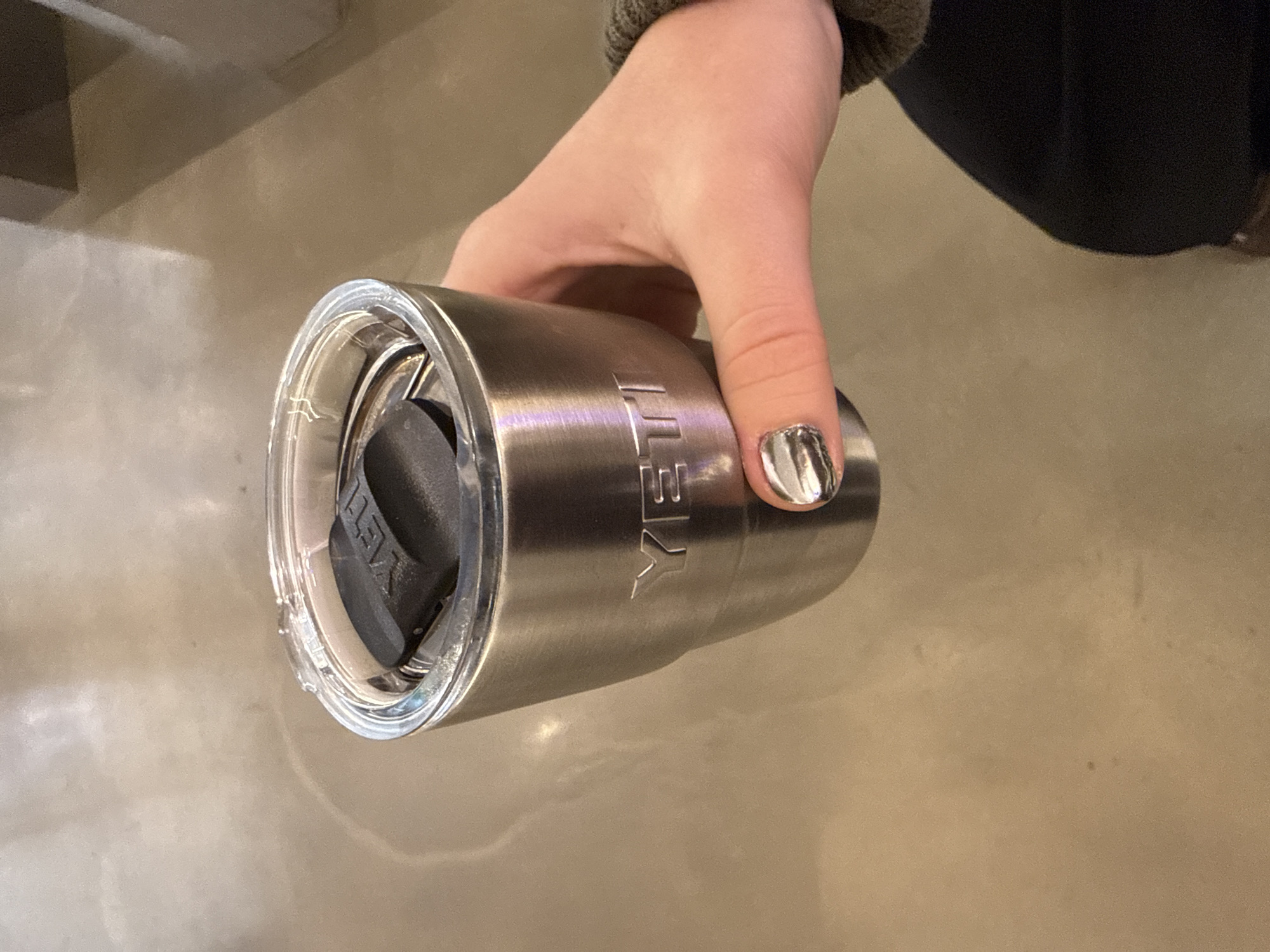 A silver Yeti tumbler is being held in a person's hand with silver nail polish. The background is a neutral beige color and appears to be indoors. There is no evidence of London, United Kingdom.