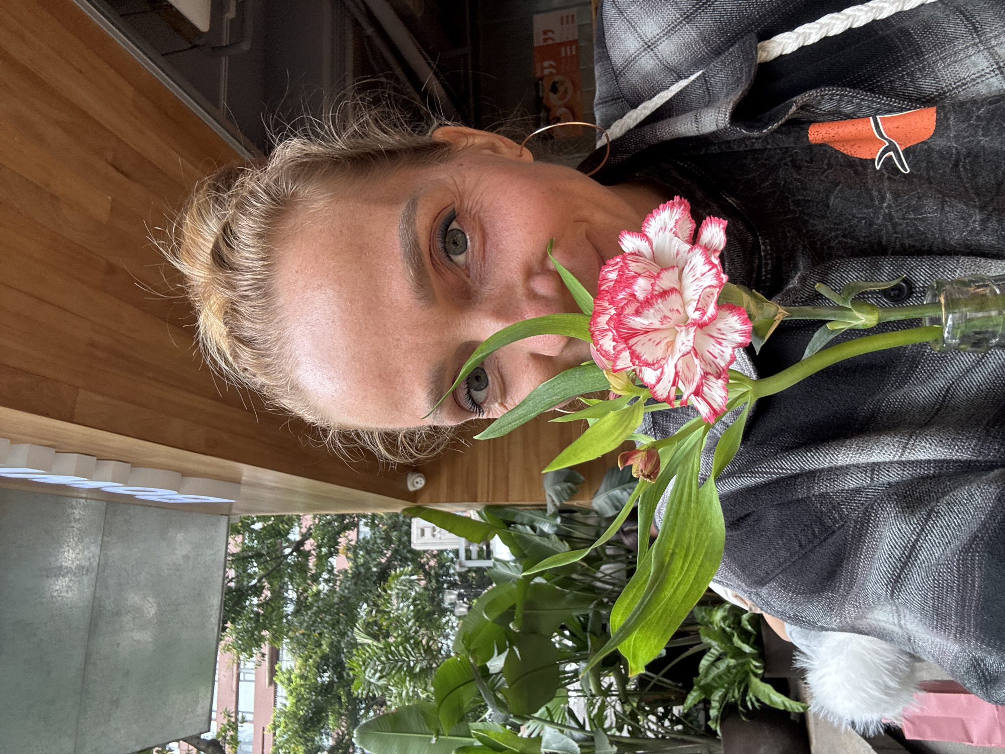 A rotated close-up captures a blonde-haired woman with light skin, looking at the camera while holding a red and white carnation flower near her face. She is wearing a grey plaid shirt. The background is a mix of indoor and outdoor elements: wooden structures, greenery including banana leaves and other plants, and a glimpse of urban buildings, suggestive of an outdoor setting. The absence of explicit clues makes it challenging to confirm the location as Yuzhong District, China. The objects and plants seem to be in her hands.