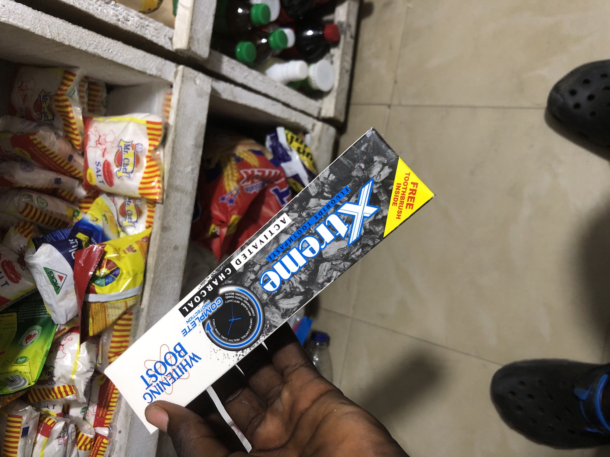 A Nigerian person's left hand, with dark skin and visible fingernails, holds a tube of "Xtreme" brand toothpaste. The toothpaste tube is predominantly white with blue and black accents, featuring the text "WHITENING BOOST COMPLETE" and "ACTIVATED CHARCOAL TOOTHPASTE". A circular graphic on the tube depicts a stylized mouth with teeth and lists benefits such as "HEALTHY GUMS", "TARTAR CONTROL", "ANTI PLAQUE", "ANTI CAVITY", "STRENGTHEN ENAMEL", and "FRESHENING BREATH". Below this, the word "Xtreme" is prominently displayed in large blue lettering over a charcoal-like texture. In the bottom left corner of the toothpaste tube, a yellow triangle shape contains the text "FREE TOOTHBRUSH INSIDE" in black lettering, oriented upside down. The background consists of shelves filled with various packaged goods, including what appears to be food items and beverages, suggesting a retail environment, possibly a small shop or market stall in Ado, Nigeria. The floor appears to be tiled, and a person's dark-colored shoe is visible in the lower left corner of the frame.
