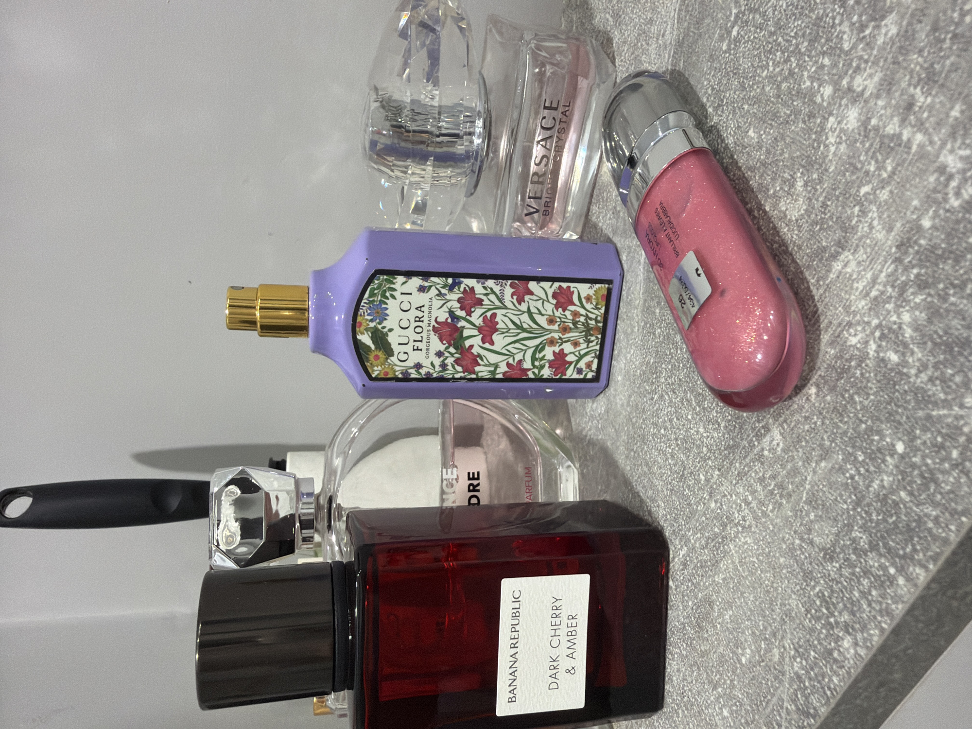 A collection of beauty and fragrance products are arranged on a textured surface, likely a countertop. Visible are: a bottle of Gucci Flora Gorgeous Magnolia perfume with a floral design and a purple bottle; a bottle of Banana Republic Dark Cherry & Amber fragrance in a rectangular, dark red bottle; a Versace Bright Crystal miniature perfume bottle; a pink lip gloss tube; and other perfume bottles with clear or crystal-shaped caps. The photo appears to be taken indoors under artificial lighting. There is no evidence to confirm the location of this scene being in the city of Ovoshchnoy, Russia.