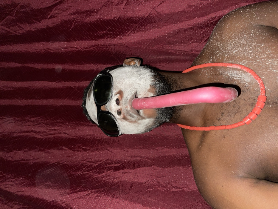 A person lies on a maroon surface, face covered in white powder and obscured by black sunglasses. A pink cylindrical object emerges from their mouth, extending towards the right. An orange beaded necklace encircles their neck and rests on their right shoulder. The person's skin is dark, with white powder also visible on their shoulder and chest. The overall composition suggests an unconventional, possibly artistic, presentation.