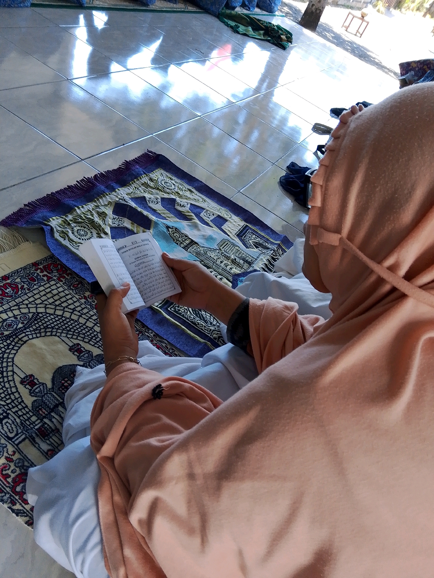 In Kuripan Utara, Indonesia, a person wearing a peach hijab and matching clothing is seated on a prayer mat, reading a book. The person is holding the book open with both hands, with their fingers supporting the pages. The prayer mat is patterned with a blue and white design and has a fringed edge. Another prayer mat with a red and blue pattern is visible beneath the first. The scene is lit by sunlight streaming in from above, creating bright reflections on the tiled floor. The environment appears to be an outdoor or semi-outdoor space, as indicated by the natural light and the presence of what seems to be a tree in the background. There are also other folded prayer mats and dark-colored footwear visible on the floor.