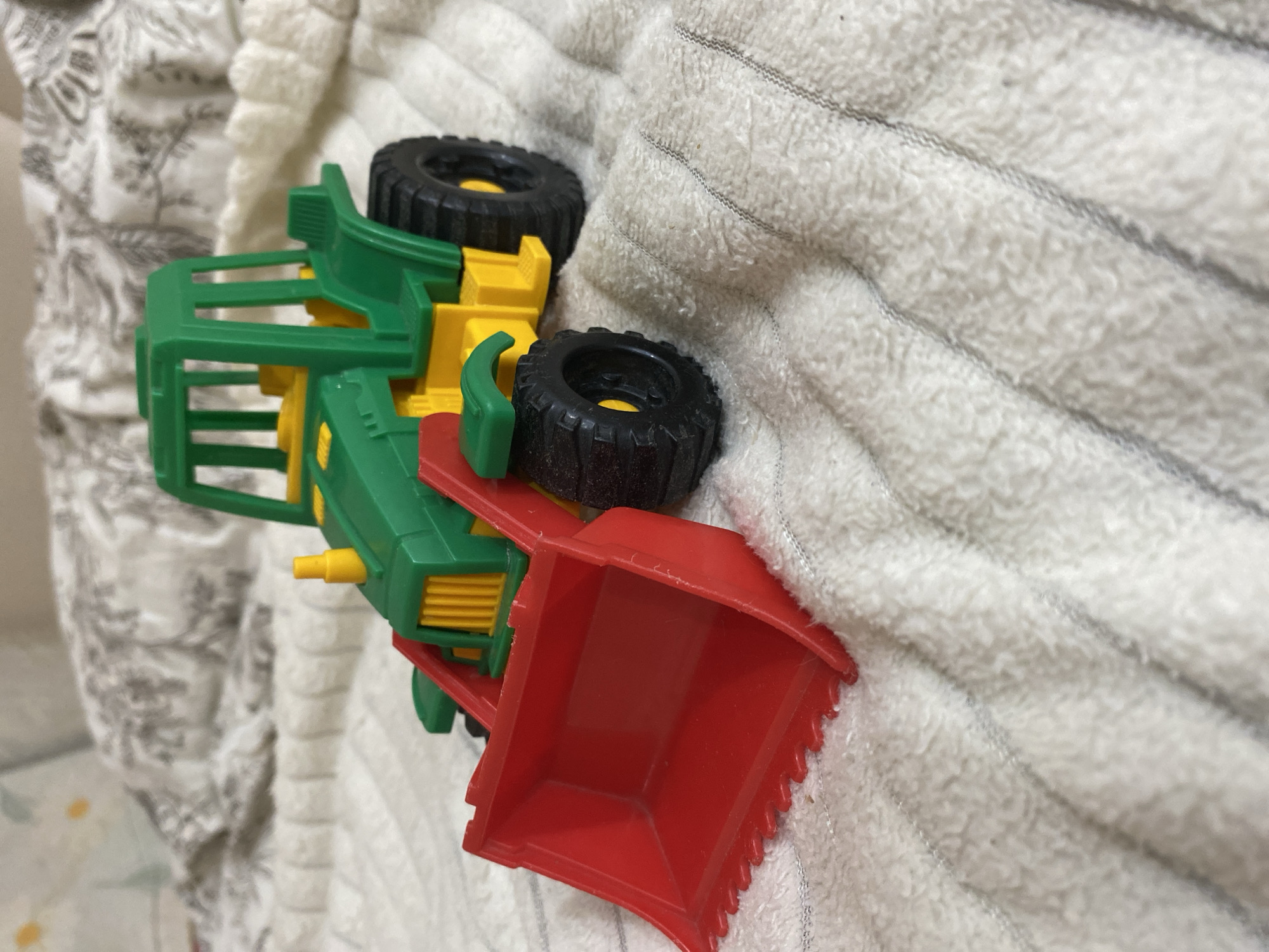 A toy front loader is placed on a plush, white blanket with visible stitched lines. The toy is primarily green and yellow, with a bright red bucket at the front. The bucket has a toothed edge. The front loader has black wheels with yellow accents. The background shows a blurred pattern of light-colored fabric, possibly a bedspread or similar textile. There are no people present, and no activity is depicted. The scene is static. The location is not discernible from the image.