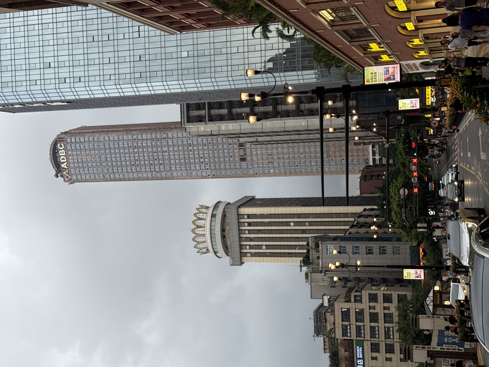 The image depicts a street scene in Yuzhong District, China, under an overcast sky. Dominating the skyline are several tall buildings. To the left, a building with a distinct crown-like structure at its apex stands prominently. In the center, a very tall, reddish-brown skyscraper with the "ADBC" logo visible near its top rises into the sky. To the right, a modern glass-fronted skyscraper reflects the cloudy sky. The street level features a mix of commercial buildings with arched windows and illuminated signage, as well as traffic lights and street lamps. Cars are visible on the road, suggesting active traffic. Pedestrians are walking along the sidewalks, some of whom are grouped together. Lush greenery can be seen interspersed between the buildings and along the street. The overall impression is of a busy urban environment in a developed district.
