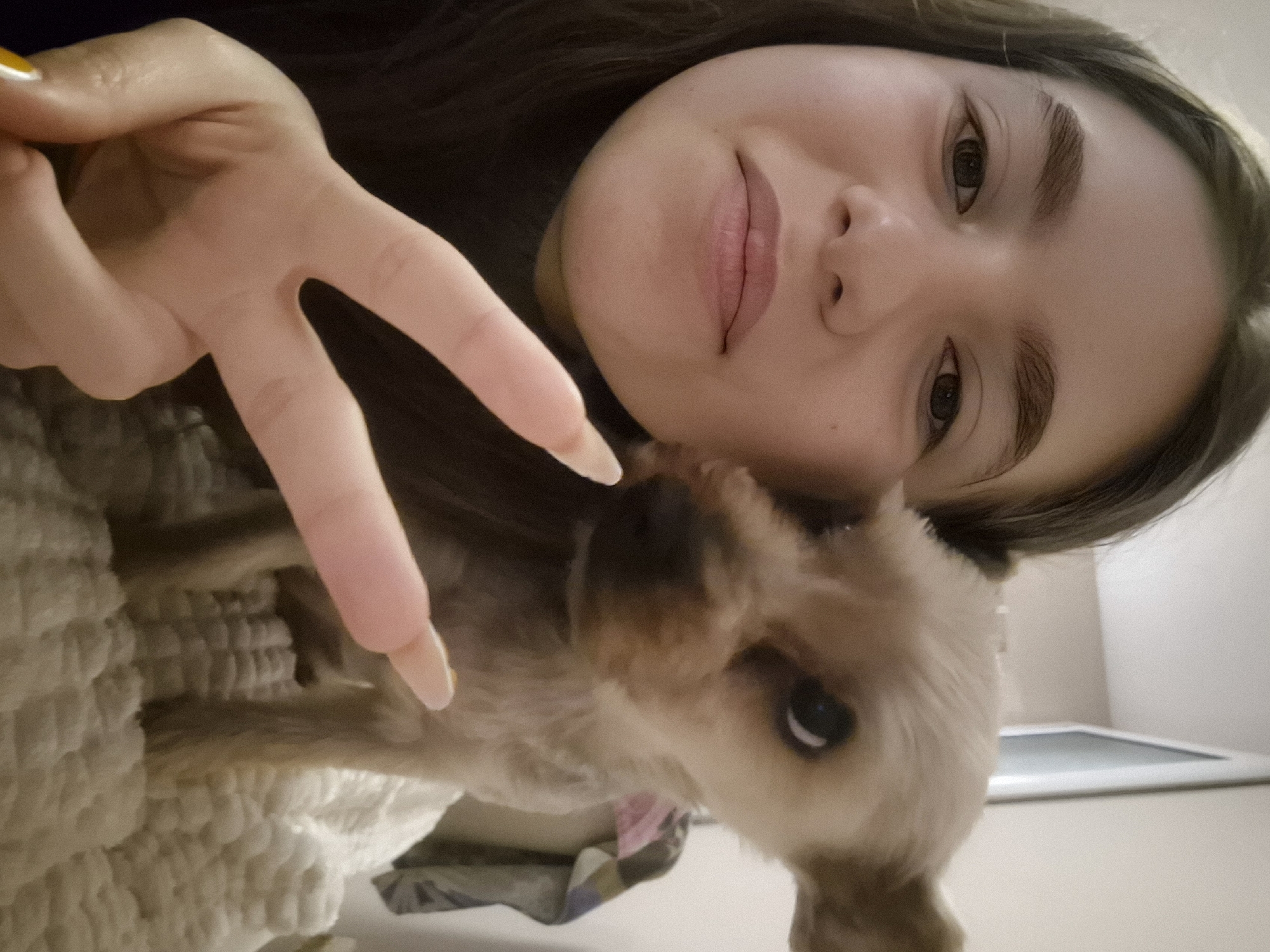 A young woman is taking a selfie with a small, light-brown dog. The woman is holding up her hand in a peace sign, with her fingers near the dog's face. The background is blurry, showing a bed with a textured blanket and a glimpse of a white wall with a monitor in the back. The location is undetermined, and there is no evidence to suggest it is in Pavlikeni, Bulgaria.