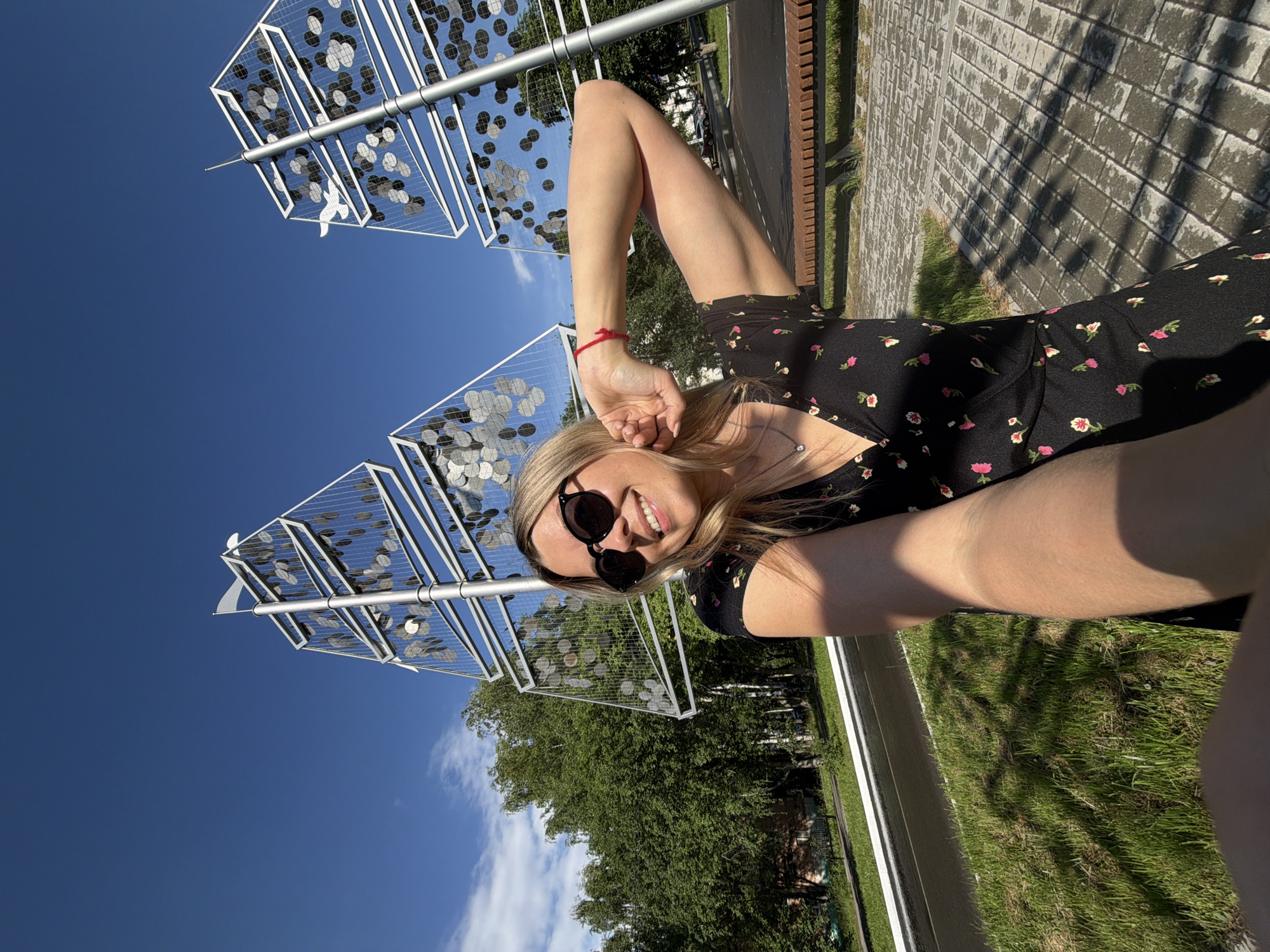 A blonde woman with sunglasses and a black floral dress poses for a selfie in a park. She is standing in front of a modern art installation that resembles sails, with a clear blue sky overhead. In the background, there are trees, a paved walkway, and a bench. The woman is smiling and has her arm raised. This scene is located in Chaikovsky, Russia.
