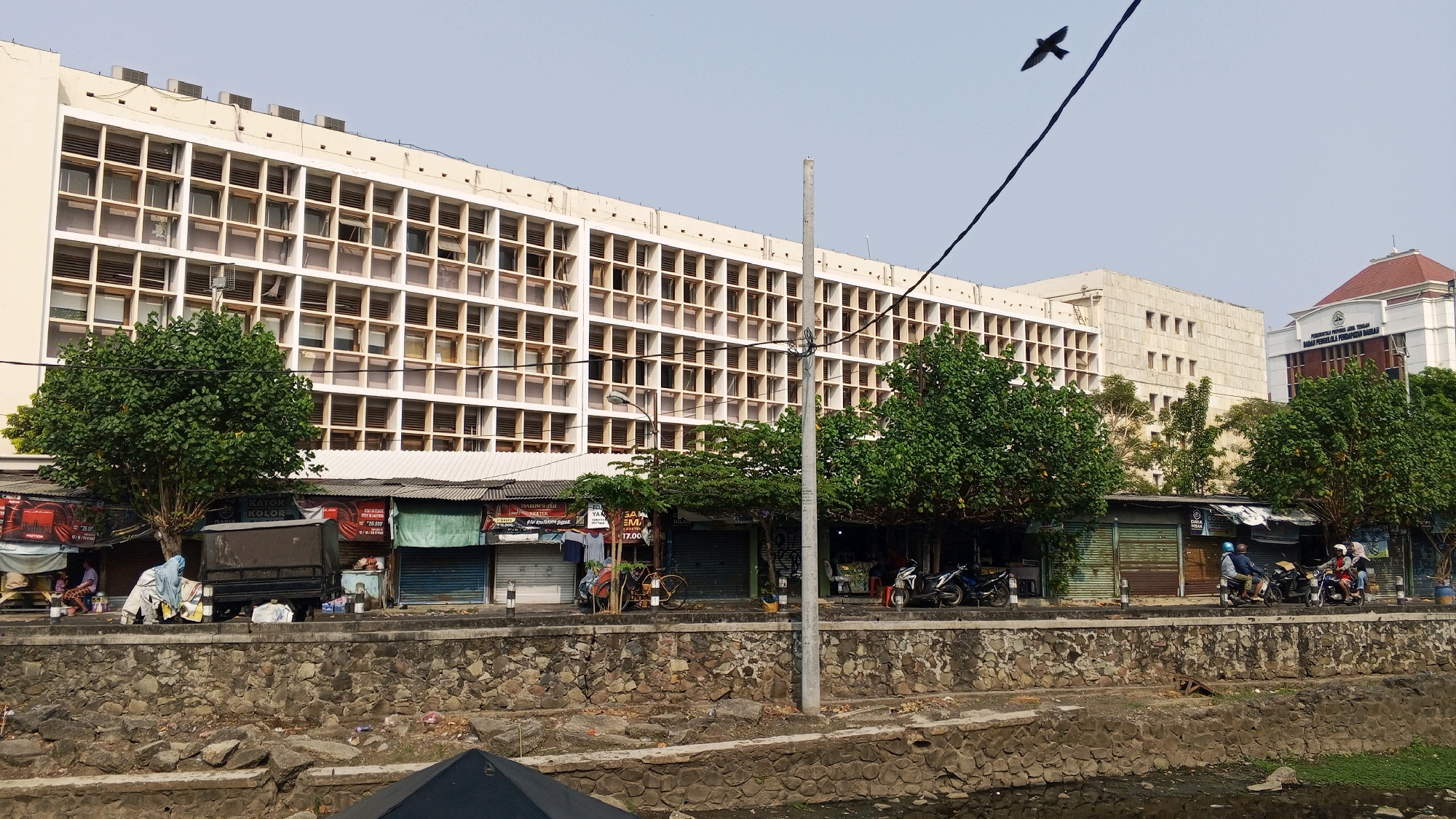 The image depicts a street scene in Semarang, Indonesia, in front of a large, multi-story building with a grid-like facade of windows. The building appears to be an institutional or office building. In the foreground, a stone-lined embankment runs along a body of water, which appears to be a canal or river. Along the embankment, a row of small shops with corrugated metal roofs are situated. Some of these shops are closed, with rolling metal shutters. Several trees are planted in front of the shops, providing shade.
Several people are present in the scene. On the left side of the image, two people are seated under an awning, one of whom appears to be a woman wearing a headscarf. A dark-colored truck is parked behind them. Further to the right, three people are riding on two motorcycles. One motorcycle is stopped, with two people on it, and another is moving, with a single rider. A bicycle is parked near one of the shops.
The sky is clear and overcast, with a single bird in flight in the upper left portion of the frame. Electrical wires are visible, stretching across the scene. In the background, another building with a more classical architectural style, featuring a prominent red-tiled roof, is visible to the right of the main building. This building has signage that reads "PEMERINTAH PROVINSI JAWA TENGAH BADAN PENGELOLA PENDAPATAN DAERAH". The overall impression is a bustling urban environment with a mix of old and new architecture, and everyday street life.