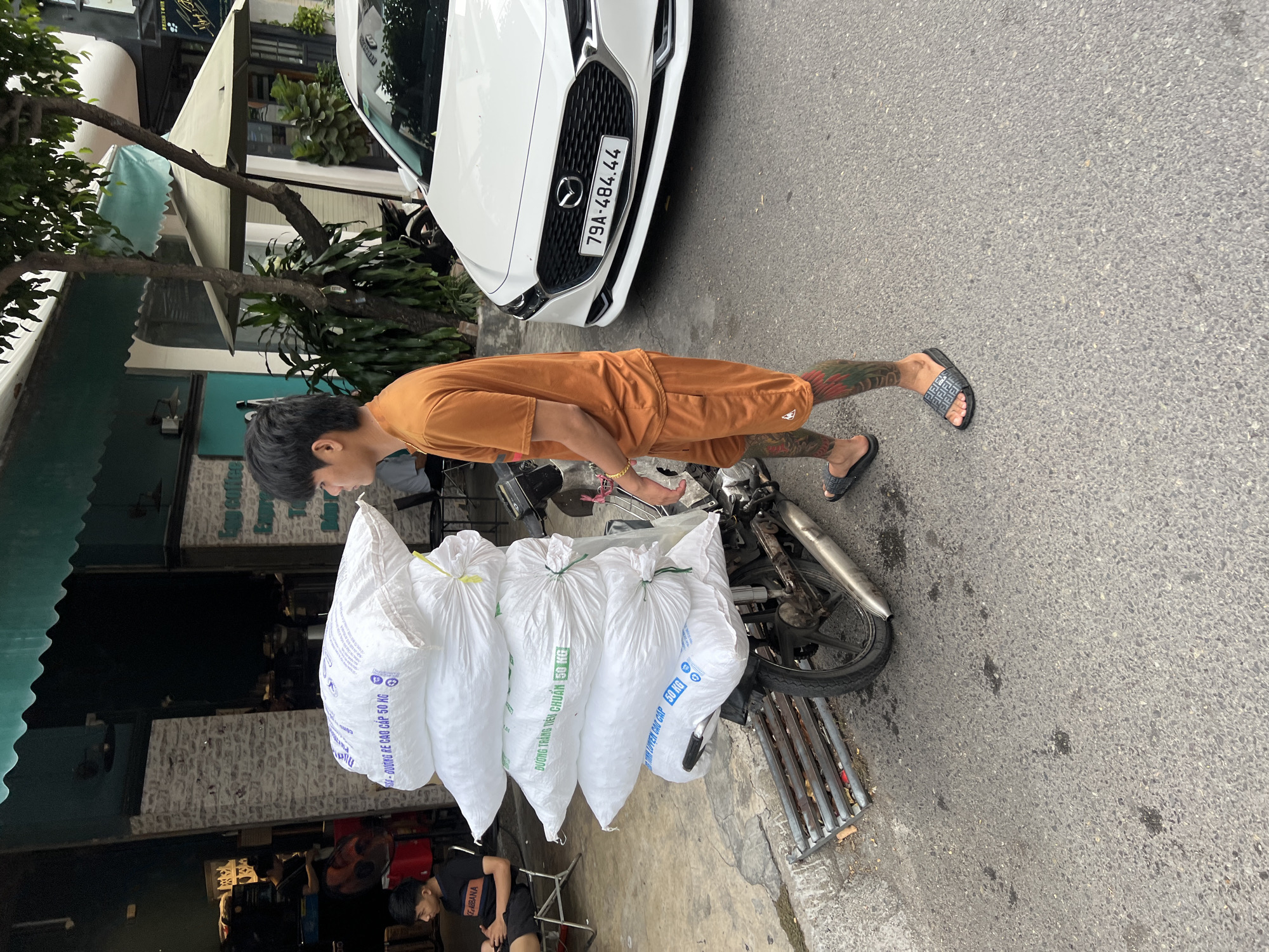 In Nha Trang, Vietnam, a young man wearing an orange t-shirt and shorts stands beside a motorcycle overloaded with five large white sacks. The sacks, likely containing goods, are stacked high on the rear of the motorcycle, with green ties securing them. The man has tattoos on his legs and is wearing black slides. The motorcycle is parked on an asphalt road, with a metal grate on the left side of the frame. In the background, a white car with a license plate reading "79A-484.44" is parked. Buildings line the street, one with a green awning and another featuring brickwork. A person can be seen sitting on a chair outside one of the buildings on the left. The scene appears to be a street in a commercial area.