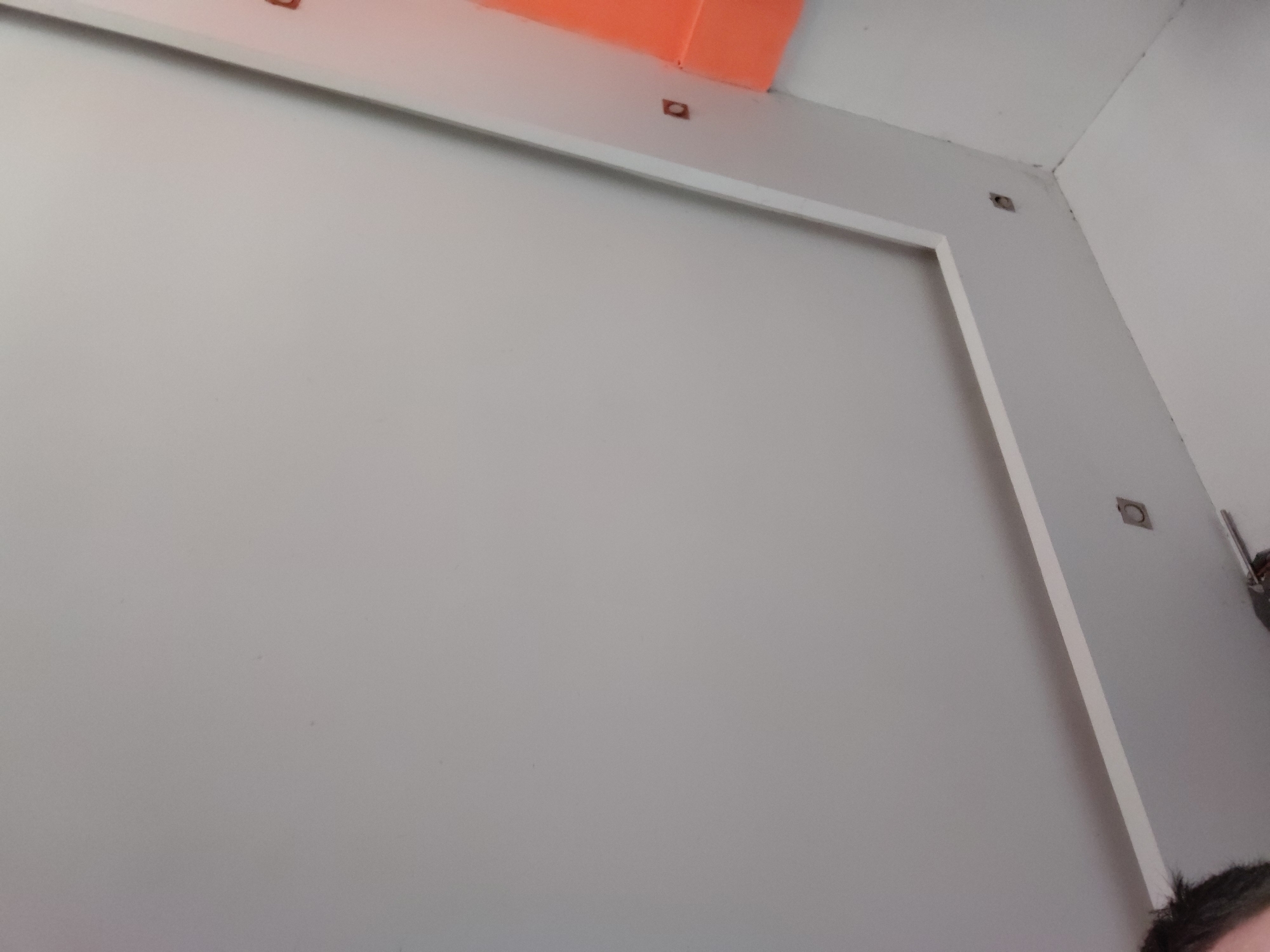 The image displays an indoor setting, likely a room under construction or renovation, dominated by a large, light grey wall. A white trim is installed on the wall, forming a right-angled frame along the top and right edges, suggesting a panel or a decorative feature. Above this white trim, near the top left, an orange material is visible, possibly insulation or part of the building's structural components.
Along the top edge of the room and extending down the right wall, there are several small, recessed square or circular openings. These appear to be mounting points for fixtures, electrical outlets, or part of a ceiling grid system. Some dark discoloration or dirt is noticeable in the top right corner where the walls meet the ceiling.
At the very bottom right corner of the frame, a small portion of a person's head, featuring dark hair, is visible. No specific activity is depicted other than the static view of the wall. The lighting appears even, suggesting indoor artificial light or diffuse natural light. There is no visible text in the image. The provided context indicates the location is Santa Ana, Venezuela.