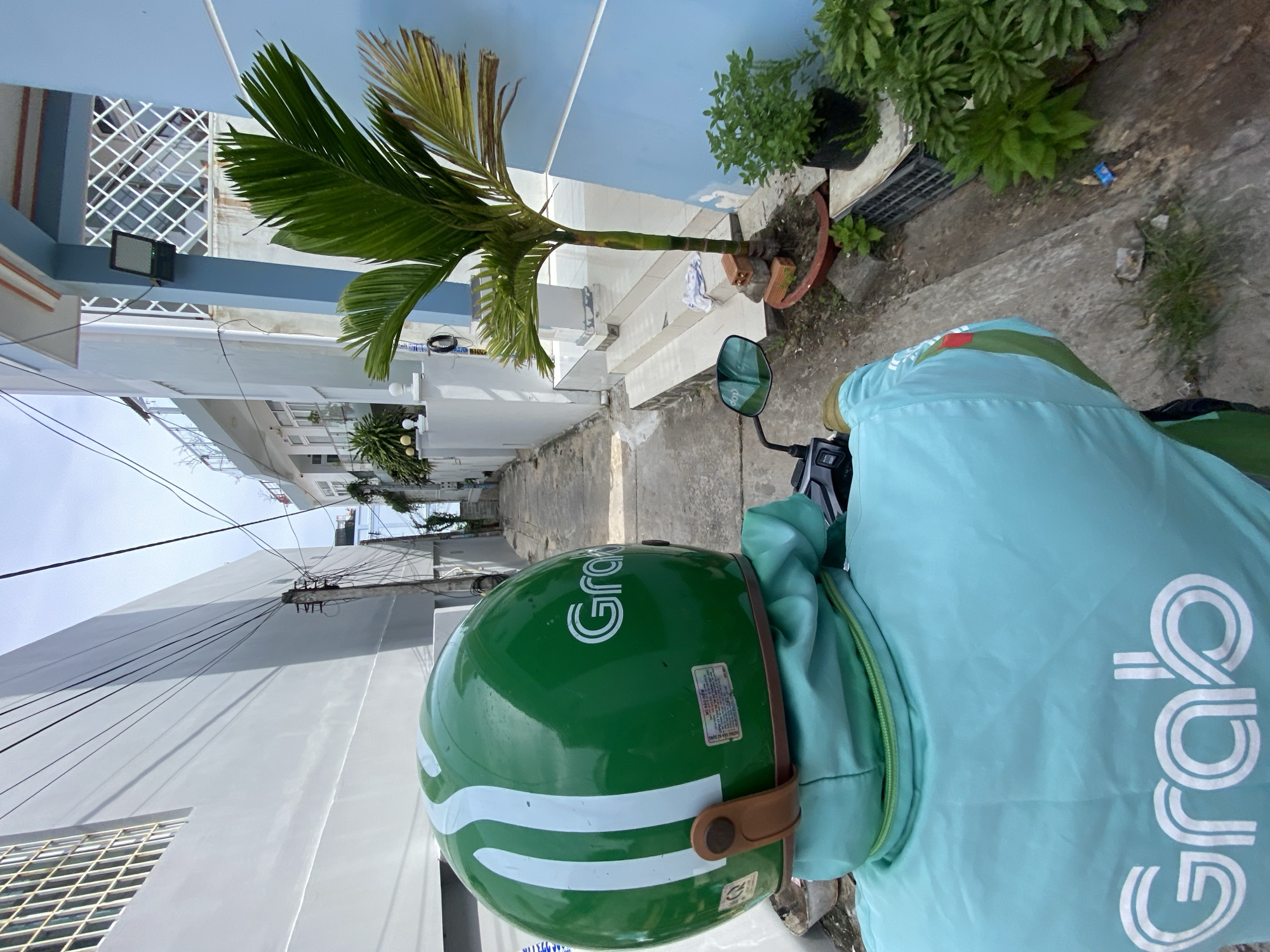 The image shows a person from behind, wearing a light blue Grab jacket and a green Grab helmet, riding a motorcycle down a narrow alleyway in Phu Thuy Ward, Vietnam. The alley is flanked by buildings, one of which is painted light blue and has a small palm tree in front of it. The ground is paved with concrete. The sky is overcast.
