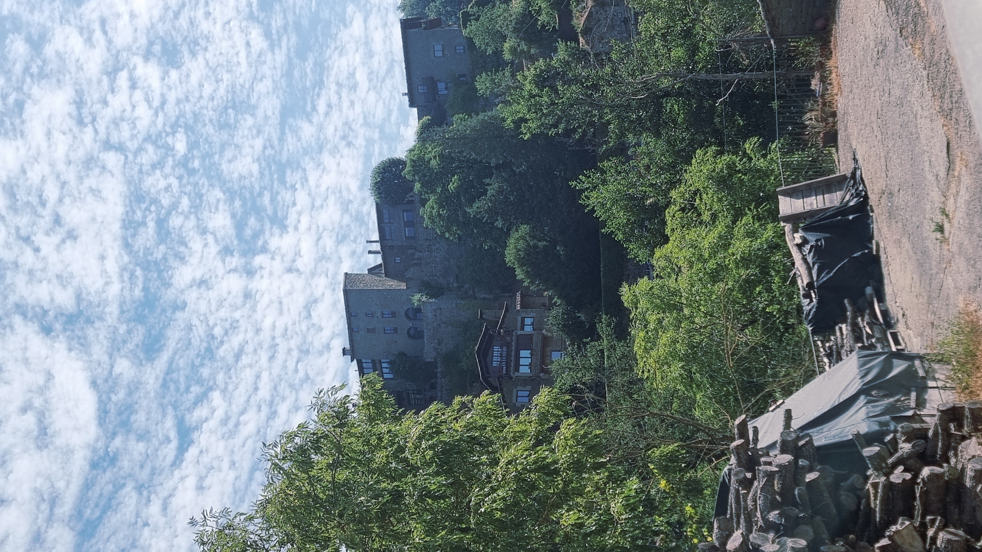 The image depicts a view of the medieval town of Rupit, Spain, nestled on a hillside under a sky filled with scattered white clouds. In the foreground, stacks of cut logs are visible, some covered with dark tarpaulin. A paved area with some scattered dry grass leads towards the base of the hill.
The middle ground features lush green trees and foliage climbing the hillside. Peeking through the trees are several stone buildings with dark-framed windows and tiled roofs, characteristic of the town's architecture. One prominent building, with a balcony and several windows, is situated lower on the slope.
Higher up the hill, larger stone structures of the town are visible, built into the slope with multiple levels and windows. These buildings appear to be made of stone blocks and have a rustic, historical appearance. The overall impression is one of an old, well-preserved village set against a natural, green landscape.