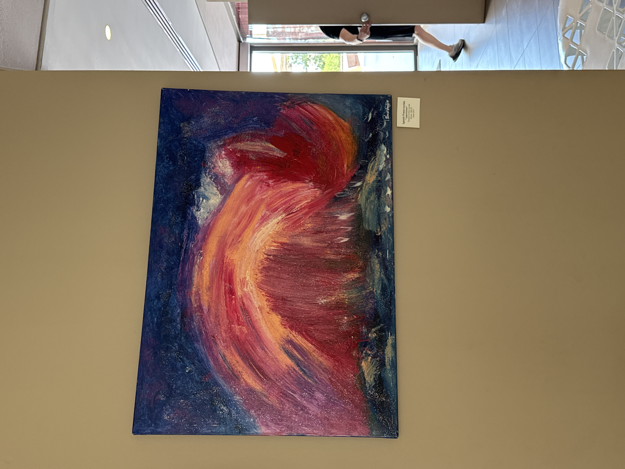 An abstract painting is prominently displayed on a plain, light beige wall. The artwork, a vertical canvas, features a vibrant, swirling form in shades of red, orange, and pink, suggesting a dynamic, almost fiery movement. These warm colors are set against a textured dark blue background, with hints of lighter blue and white brushstrokes adding to the sense of motion.
To the right of the painting, a white information card provides details about the artwork. It identifies the artist as "Ignazio Premarvecchio" and the title as "Apertura" (Opening). The medium is listed as "Tecnica Mista su tela" (Mixed technique on canvas), with dimensions of "70cm x 50cm," and the year of creation as "Anno 2017." A signature, matching the artist's name, is also visible on the upper right corner of the painting.
Above the wall, the scene reveals a bright indoor setting, likely a hallway or open space. A large glass partition or doorway provides a view of outdoor greenery and lets in ample light. The lower half of a person, wearing dark trousers and shoes, is visible as they walk past. Another person's arm, holding a cup, is also partially seen near the glass. The floor in this upper section is covered with light-colored tiles, and part of a white, open-weave chair or table is visible in the upper right corner. The overall setting suggests a public venue, such as a gallery or a building with exhibition space, located in Isola delle Femmine, Italy.
