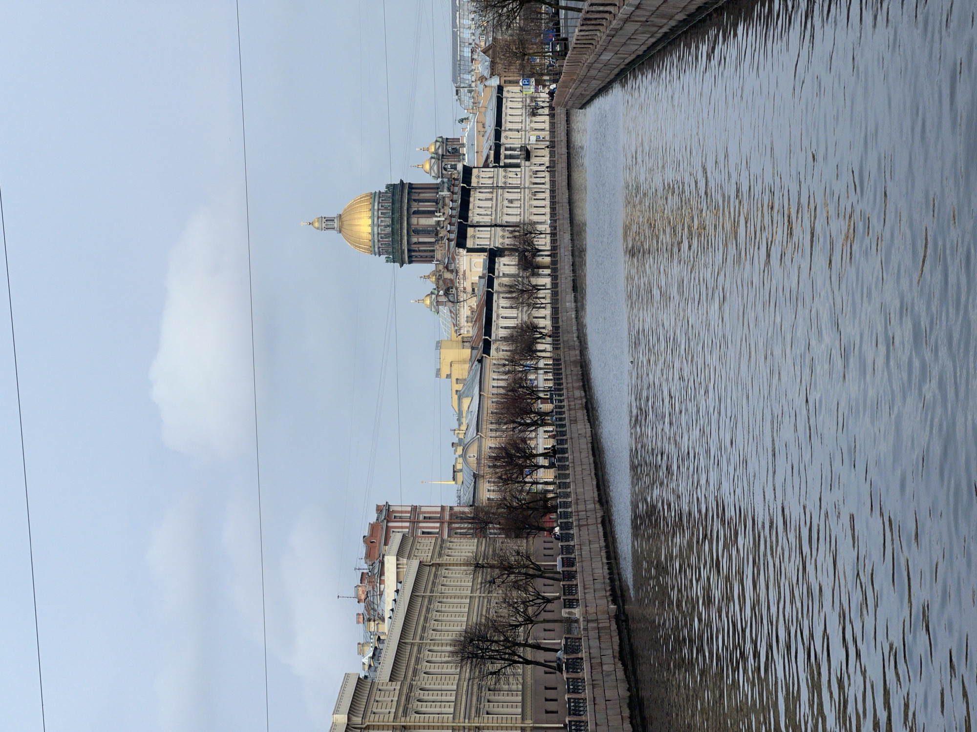 The image depicts a canal in Saint Petersburg, Russia, with St. Isaac's Cathedral visible in the background. The canal's water surface shows ripples and reflections. Buildings line the canal, with their architectural details visible. Trees, largely bare, are situated along the canal's edge. Overhead wires are seen against the overcast sky.