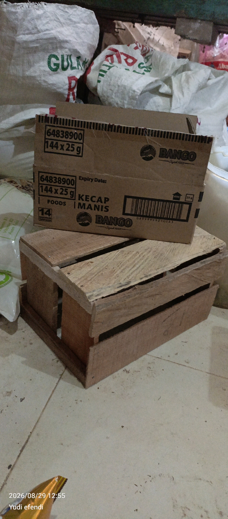The image depicts an indoor scene with stacked cardboard boxes of "Bango Kecap Manis" (sweet soy sauce) and a wooden crate. The boxes are brown cardboard, one on top of the other, with product labels indicating "64838900 144 x 25 g". The lower box also features a barcode and the "Bango" logo. In the background, partially visible white sacks with green lettering, possibly containing sugar ("GULA") and other goods, are stacked. The floor appears to be tiled and somewhat dusty. To the left, part of a white plastic bag is visible. The overall setting suggests a storage area or a retail backroom. The timestamp in the lower left corner indicates the photo was taken on 2026/08/29 at 12:55, attributed to "Yudi efendi". No specific location in Rawajitu Selatan, Indonesia is visually evident from the image content.