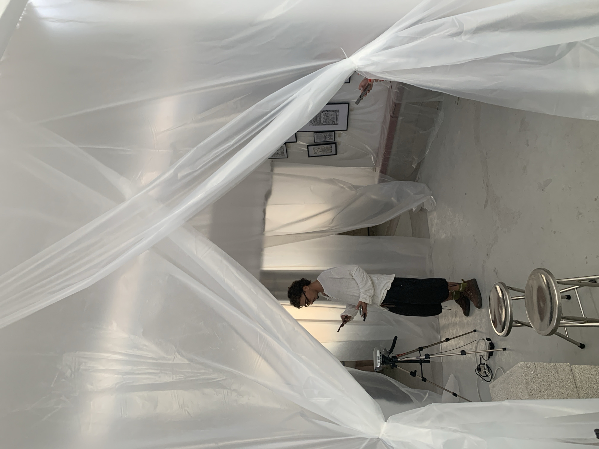 The media file captures an indoor scene, likely in Tel-Aviv, Israel, dominated by translucent white plastic sheeting that forms an enclosed or semi-enclosed space. The sheeting creates a tunnel-like structure, with folds and creases visible throughout.
At the center-right of the image, an adult individual with curly dark hair and glasses is visible. They are wearing a light-colored long-sleeved shirt and dark pants or shorts. The person is leaning forward, looking down intently at something they are holding in both hands, possibly a mobile phone and another small device or tool. Their posture suggests focus on a task.
In the background, visible through and behind the plastic sheeting, several framed artworks or pictures are displayed on a white wall. These appear to be black and white drawings or prints, arranged in a gallery-like fashion.
On the floor in the foreground, to the right of the person, there are two metallic, round-seated stools. To the left of the stools, a camera tripod is set up with a device mounted on top, possibly a camera or light source, and some electrical cables are visible on the light-colored floor. The walls and floor are plain and light-colored, contributing to a minimalist and temporary aesthetic. Faint red structural elements can be seen behind the plastic sheeting in the far background.
The overall scene suggests the preparation or setup of an art installation, exhibition, or a work environment within a larger space, with the plastic sheeting creating a distinct area for the activity. No discernible text is visible on any objects or surfaces.