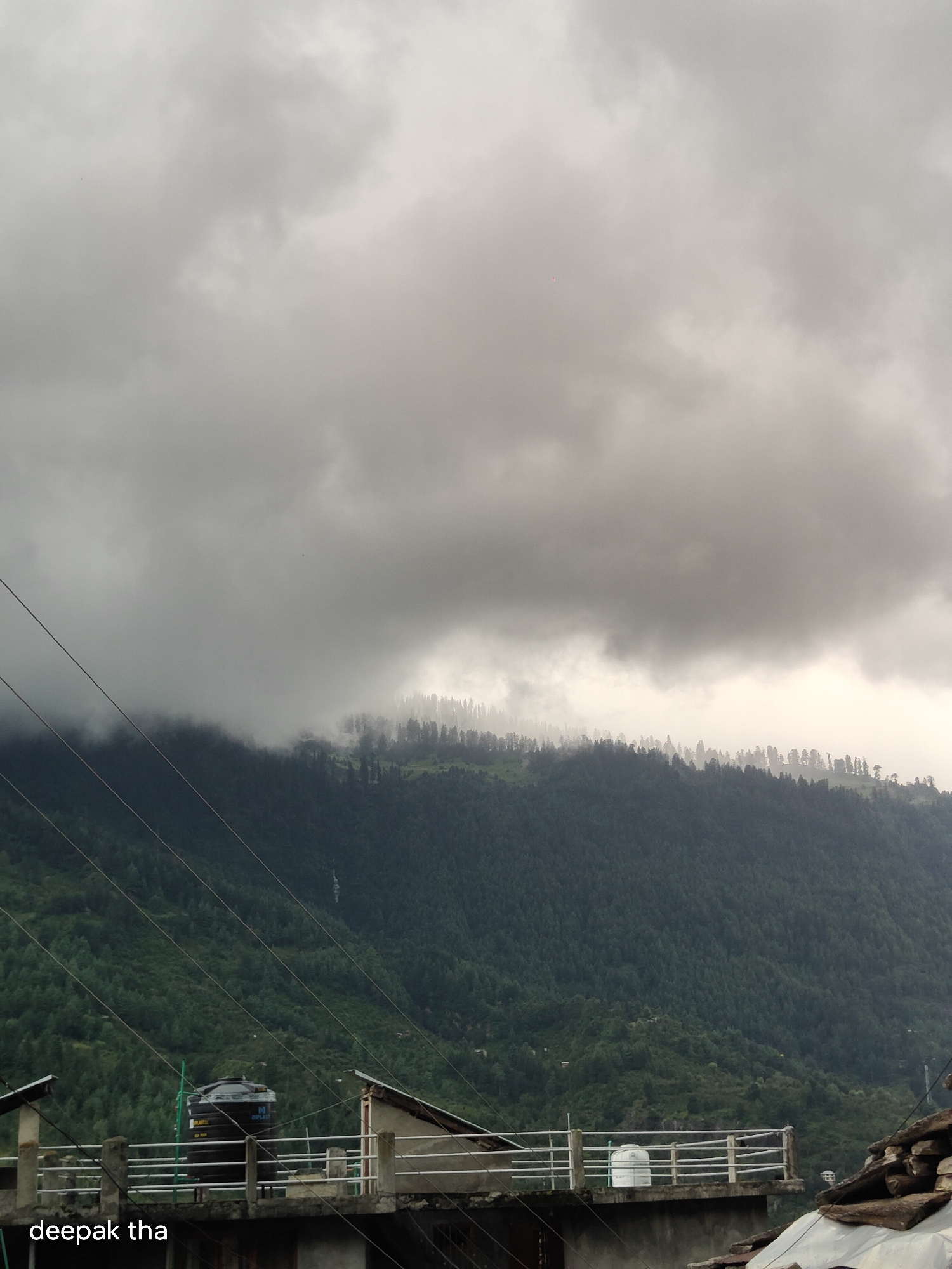 The image captures a cloudy sky over a densely forested mountain range. In the foreground, a rooftop structure is visible with a black water tank and a white water tank. There are also power lines crossing the frame diagonally. The text "deepak tha" is superimposed in the lower left corner of the image. The scene appears to be of a mountainous region in Vashisht, India, likely taken during a period of overcast weather.