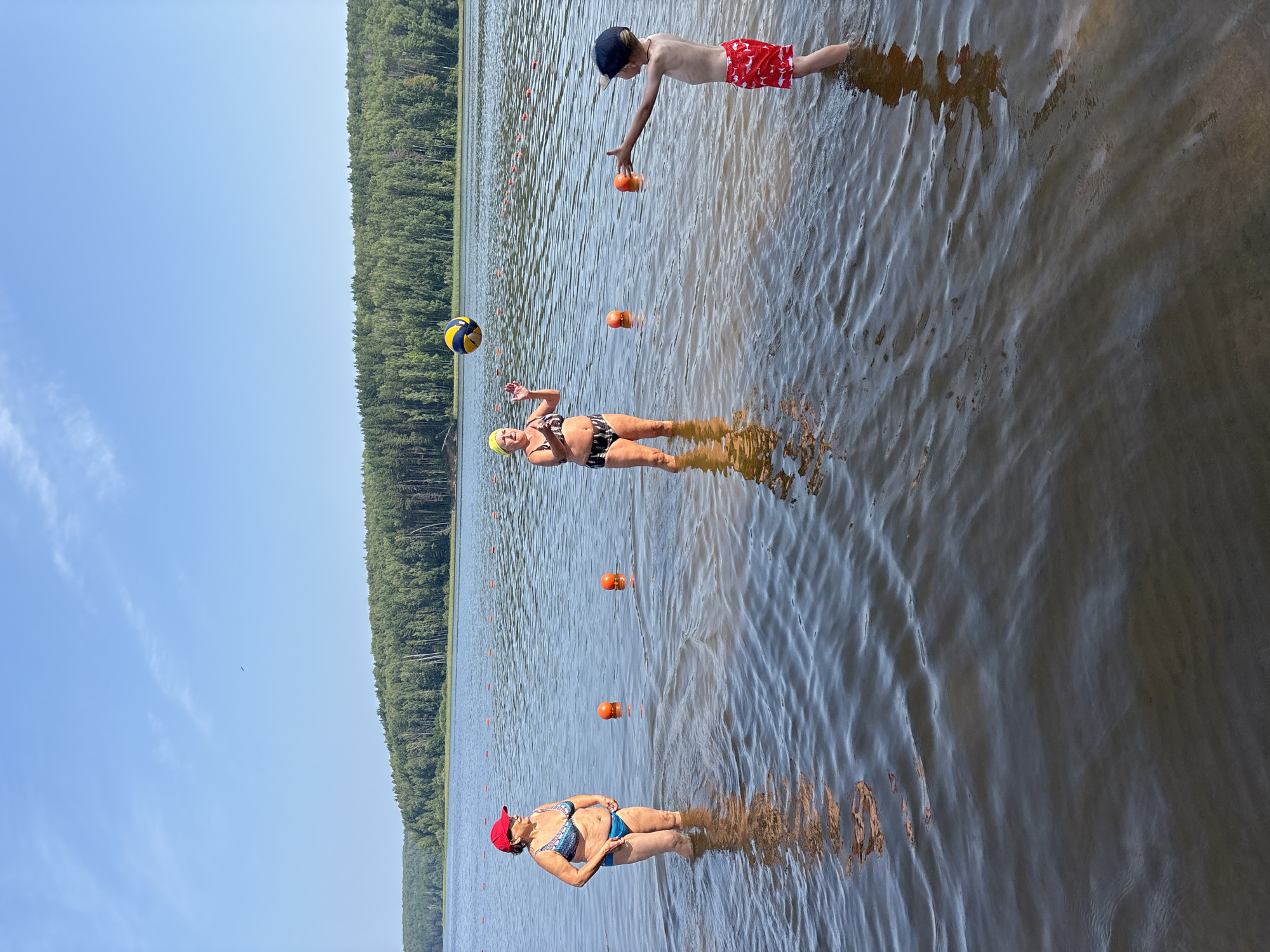 A wide shot captures a group of people wading in a lake on a sunny day. The scene is oriented vertically, with the water in the foreground and a dense, green forest bordering the lake in the background. The sky above is a clear, bright blue with a few wispy clouds.
Three people are visible in the water. In the foreground, a woman wearing a red cap and a patterned bikini stands in the shallow water, looking to her left. Further out in the lake, a woman in a black and white bikini and a yellow swim cap is positioned in the water, with a volleyball floating near her. She appears to be in the process of playing or interacting with the ball. In the background, a young boy wearing red patterned swim trunks and a dark cap is in the water, reaching out towards two orange buoys.
Orange buoys are scattered across the lake, marking a swimming area or lane. The water is rippled, reflecting the sunlight and the surrounding environment. The overall mood is relaxed and recreational, suggesting a summer day at a lake. The lighting indicates it is daytime, likely midday or early afternoon, given the brightness of the sun. The presence of buoys, a volleyball, and swim attire points to a recreational activity, possibly swimming or water games. No text is visible in the image. The location context provided is Chaykovsky, Russia.