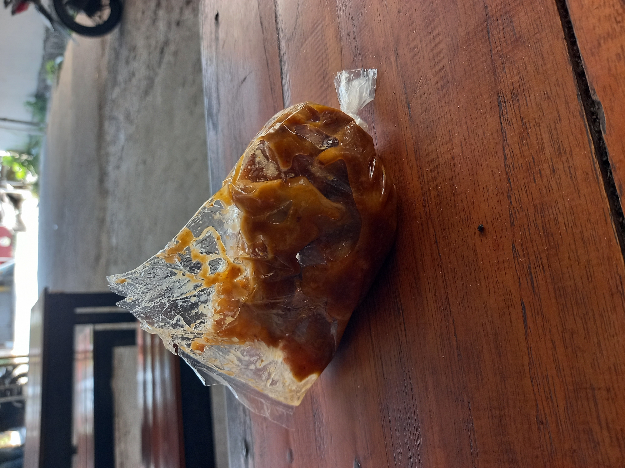 A plastic bag containing food, possibly meat in a sauce, sits on a wooden table. The bag is tied with a twist tie. In the background, out of focus, are tables and chairs, suggesting an outdoor eating area. A motorcycle is partially visible on the right side of the image. The location is identified as Pelemwatu, Indonesia. The scene appears to be a casual outdoor dining setting.