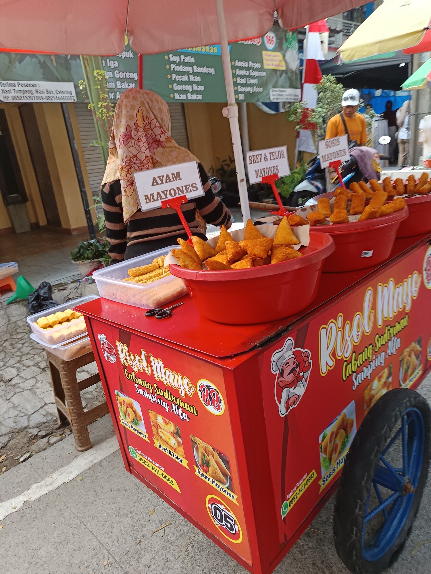 An outdoor food cart is set up on a cracked sidewalk in Sodong, Indonesia, with a bright red cart as the central focus. The cart is adorned with images of food and text that appears to be in Indonesian. Several containers of fried food are displayed in red bowls on top of the cart. To the left of the cart, there is a small brown stool with plastic containers of food. Behind the cart, a person wearing a headscarf is seen from the back, interacting with a sign that reads "AYAM MAYONES". Other signs behind the food cart advertise different food items. The scene is framed by umbrellas that provide shade.