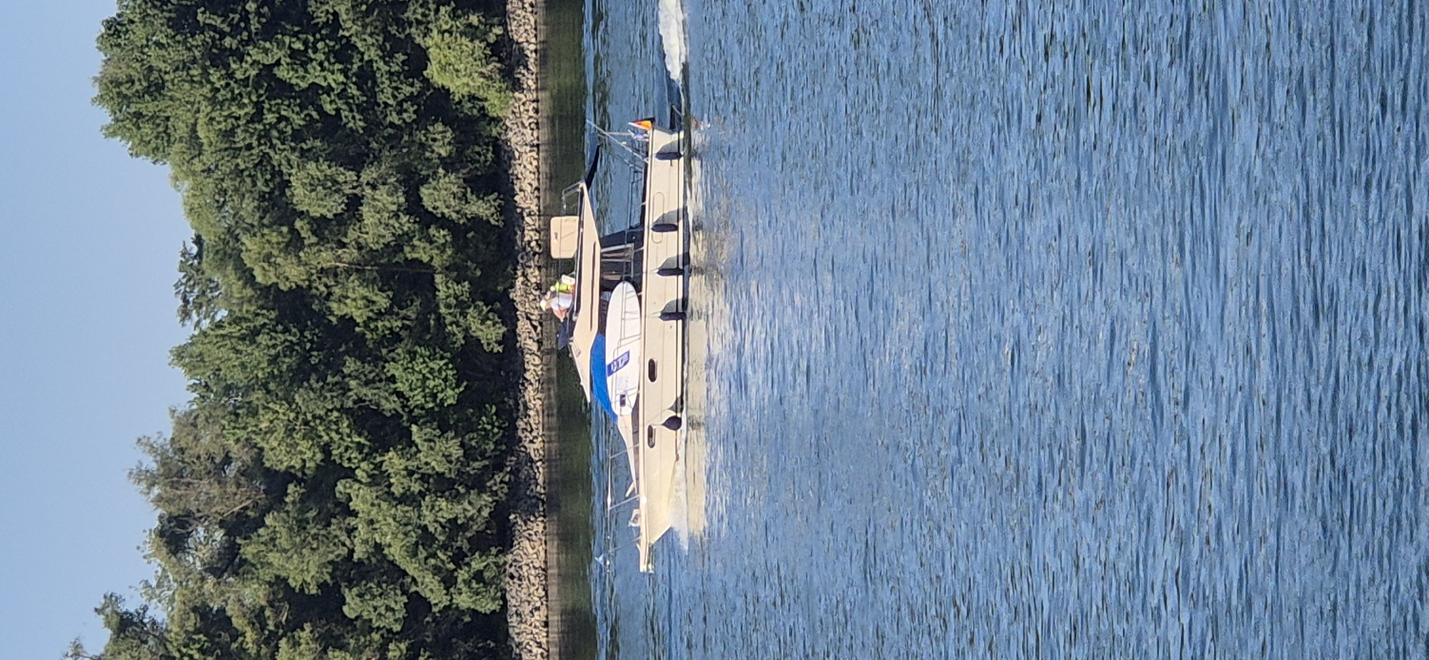 A beige motorboat with a blue canopy is traveling on a river. Two people are visible on the upper deck of the boat. The boat is leaving a wake behind it. On the left side of the boat, a white surfboard is secured vertically. The river is calm and reflects the blue sky. The background features a dense line of green trees and a rocky embankment. The scene is set on the Rhine River in Mainz, Germany. The sky is clear and blue.