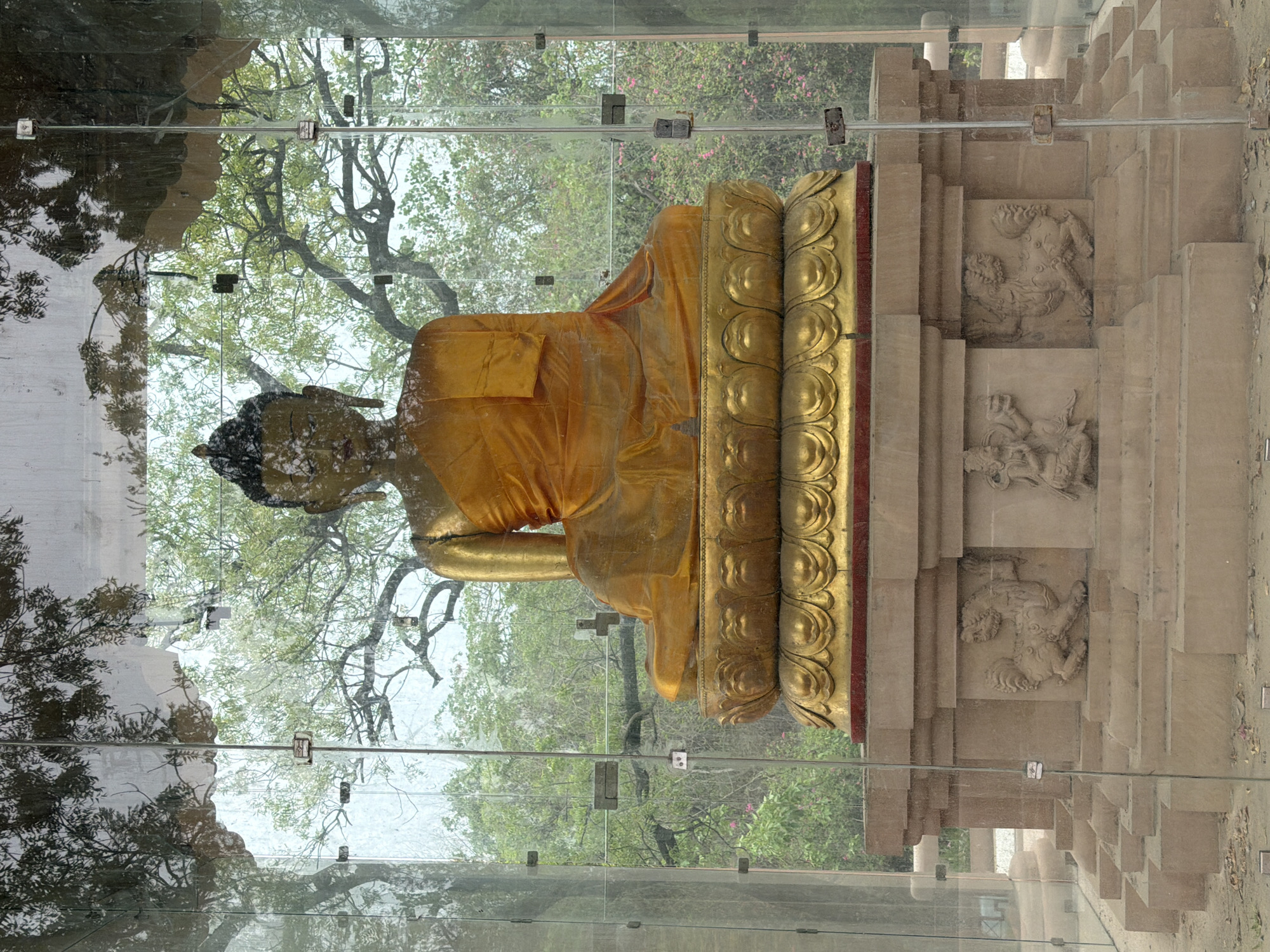 A golden Buddha statue sits on a tiered base within a glass enclosure in New Delhi, India. The statue is seated in a meditative pose, with its right hand resting on its lap and its left hand in its lap. The statue is adorned with a saffron robe. The base of the statue is decorated with intricate carvings of lions and other mythical creatures. The glass enclosure is surrounded by trees and foliage, suggesting an outdoor setting. The overall scene conveys a sense of peace and tranquility.