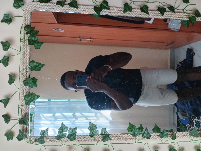 A person is reflected in a mirror decorated with rope and faux ivy. The individual is taking a photo with a blue phone. They wear a black shirt and light-colored shorts. The reflection shows a bedroom with a light brown dresser in the background. Below the mirror, a window is partially visible. A bed and shoes are seen in the lower right corner. The room has neutral-colored walls.