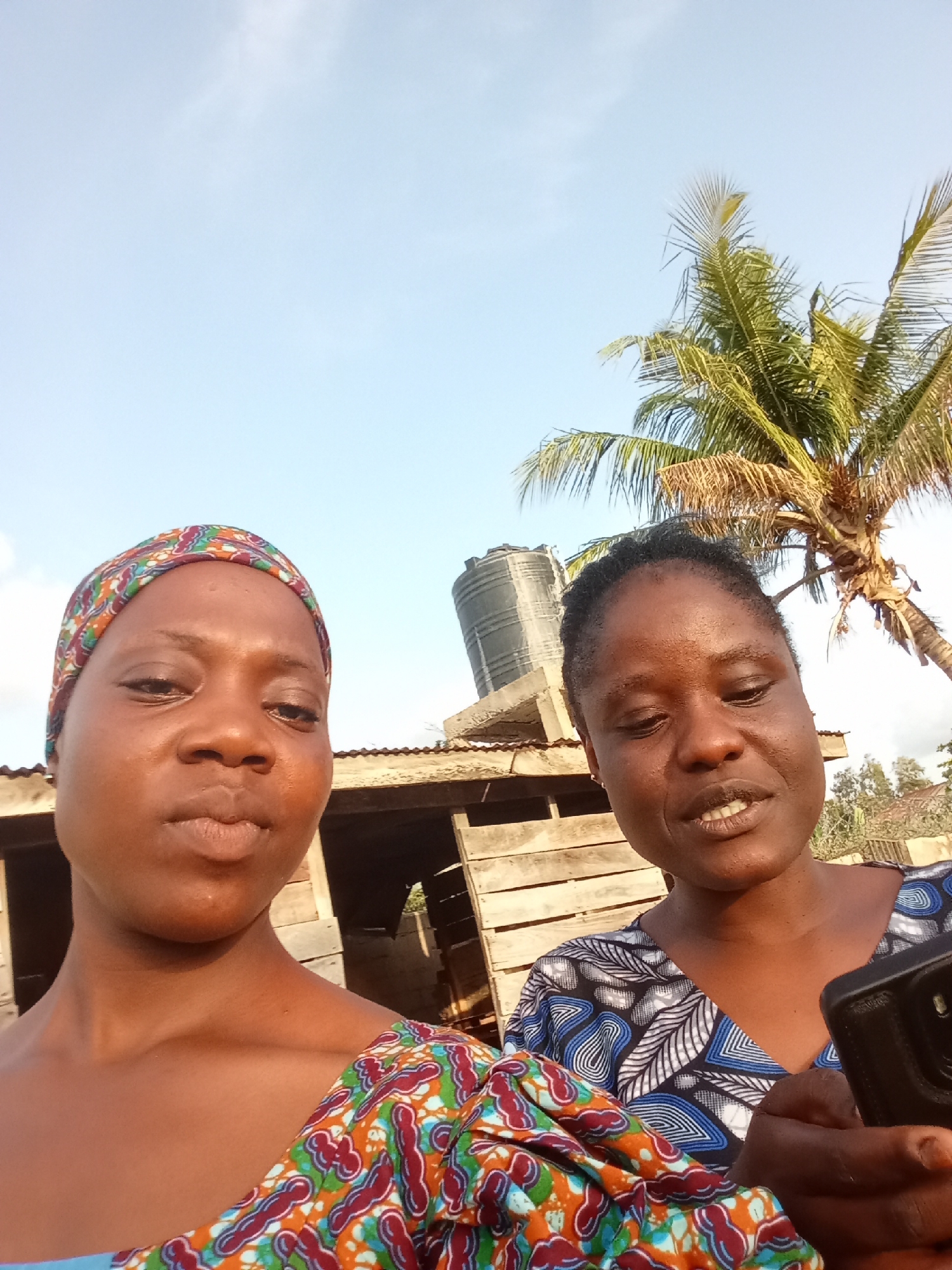 Two women are captured in a close-up selfie, with a background that suggests an outdoor, possibly rural, setting. The woman on the left is wearing a colorful headwrap with patterns of red, blue, green, and orange. Her gaze is directed towards the camera. The woman on the right has her hair pulled back and is wearing a blue and white patterned top. She is holding a black smartphone in her right hand, seemingly taking the photo. Her expression is animated, with her mouth slightly open as if speaking.
Behind the women, a rustic wooden structure is visible, possibly a shed or a partially built dwelling. To the right of the structure, a large black water tank is mounted on a concrete base. Further in the background, a palm tree with vibrant green fronds stands tall against a clear, bright blue sky, indicating it is daytime. The presence of the palm tree and the style of the building suggest a tropical or sub-tropical location. The lighting is bright, with the sun casting a warm glow on the subjects' faces, hinting at a sunny day. The overall mood of the image appears relaxed and informal, likely a casual moment captured between friends or acquaintances. No visible text is present in the image.
