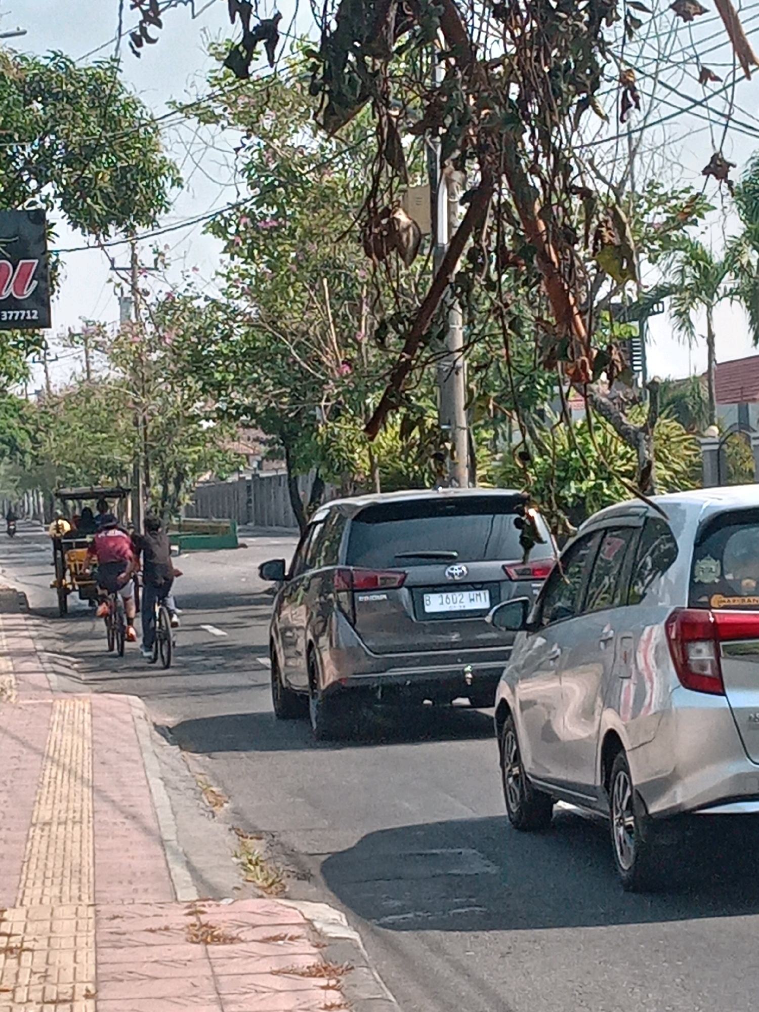 The image depicts a street scene in Yogyakarta, Indonesia. The foreground shows a sidewalk with tactile paving, typical for guiding visually impaired pedestrians. The sidewalk is bordered by a street with several vehicles. Two individuals are riding bicycles on the street, one wearing a maroon shirt and the other a dark shirt. Behind them, a motorized rickshaw, known locally as a "becak," is visible with passengers. Further down the street, cars are present, including a dark grey SUV with the license plate "B 1602 HMI" and a silver car. The background features trees, buildings, and a clear sky, suggesting an urban environment during daylight hours. The presence of the becak and the specific type of tactile paving strongly indicate a location within Indonesia, likely a city like Yogyakarta, which is known for its becak transportation and urban development.