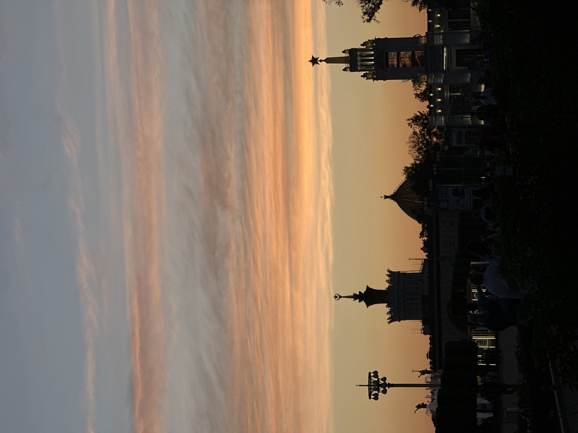 The image shows a sunset view in an urban location. The sky is filled with long, streaky clouds colored in shades of peach, orange, and light blue. Silhouetted against the sky are various architectural structures, including buildings with spires, domes, and rooftop details. One building has a prominent star on top. A pole-mounted lighting structure is also visible. Dark foliage and indistinct figures of people are present in the foreground. The location is in the city of Unknown, Unknown.