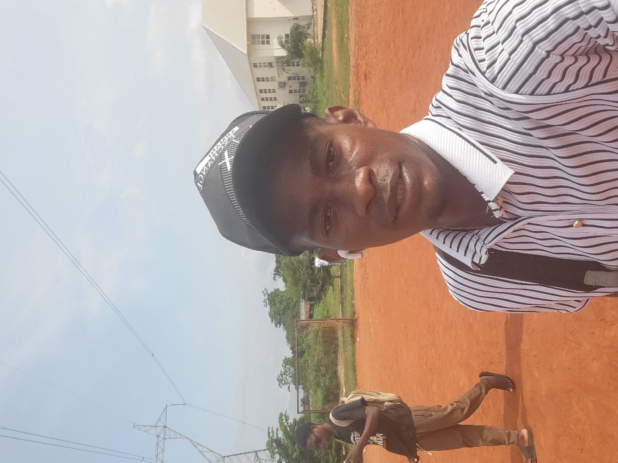 The image shows a close-up selfie of a man wearing a black baseball cap and a striped shirt. He has a light skin tone and is wearing white earbuds. In the background, there is another person wearing a black t-shirt and brown pants, carrying a backpack. They are walking on a reddish-brown dirt surface, possibly a sports field. Behind them, there are trees, a small building with a goalpost, and a large white building with many windows. Electrical pylons and power lines are visible in the sky. The scene appears to be an outdoor area in a semi-urban or rural setting. The location is identified as Ukukwa, Nigeria.