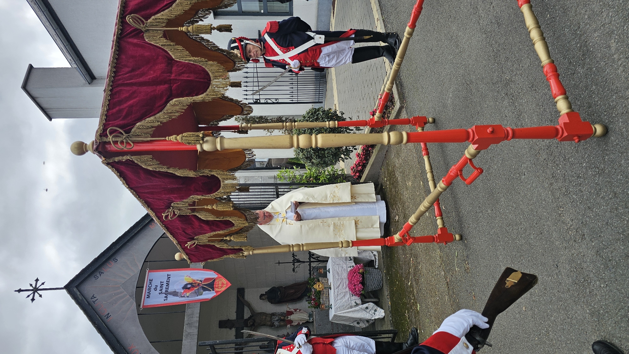 This image depicts an outdoor religious ceremony in Boussu-lez-Walcourt, Belgium, under an overcast sky. In the central foreground, a large, ornate portable ceremonial canopy with a red velvet, gold-fringed baldachin is prominent, supported by a red and gold painted wooden or metal frame.
To the left of the canopy, beneath the archway of a stone chapel or grotto, an elderly priest stands, wearing a white cope and vestments, reading from a booklet. The chapel's facade features a black cross on its roof peak and the inscription "IN SOLITA SALUS" above the arch. A vertical banner hangs from the archway, displaying "MARCHE du SAINT SACREMENT" and a smaller emblem with "16GS".
Adjacent to the priest, an outdoor altar is set up, draped with a white embroidered cloth. It holds a gold monstrance, a small bell, and arrangements of pink flowers. Inside the grotto behind the altar, a large crucifix and other religious statues are visible. Black wrought-iron gates partially enclose the chapel area.
Two uniformed guards are present. On the right side of the canopy, a guard stands at attention, wearing a dark blue coat with red lapels and cuffs, white trousers, black tall boots, and a black shako with a red plume. He holds a silver-bladed saber at his hip. A second guard, partially visible on the far left, wears a similar uniform and holds a dark brown rifle with a golden butt plate.
The ground is dark, wet pavement. Green foliage is visible in planters along the chapel's perimeter. Two faint, indistinct specks are visible against the grey sky in the upper portion of the image.