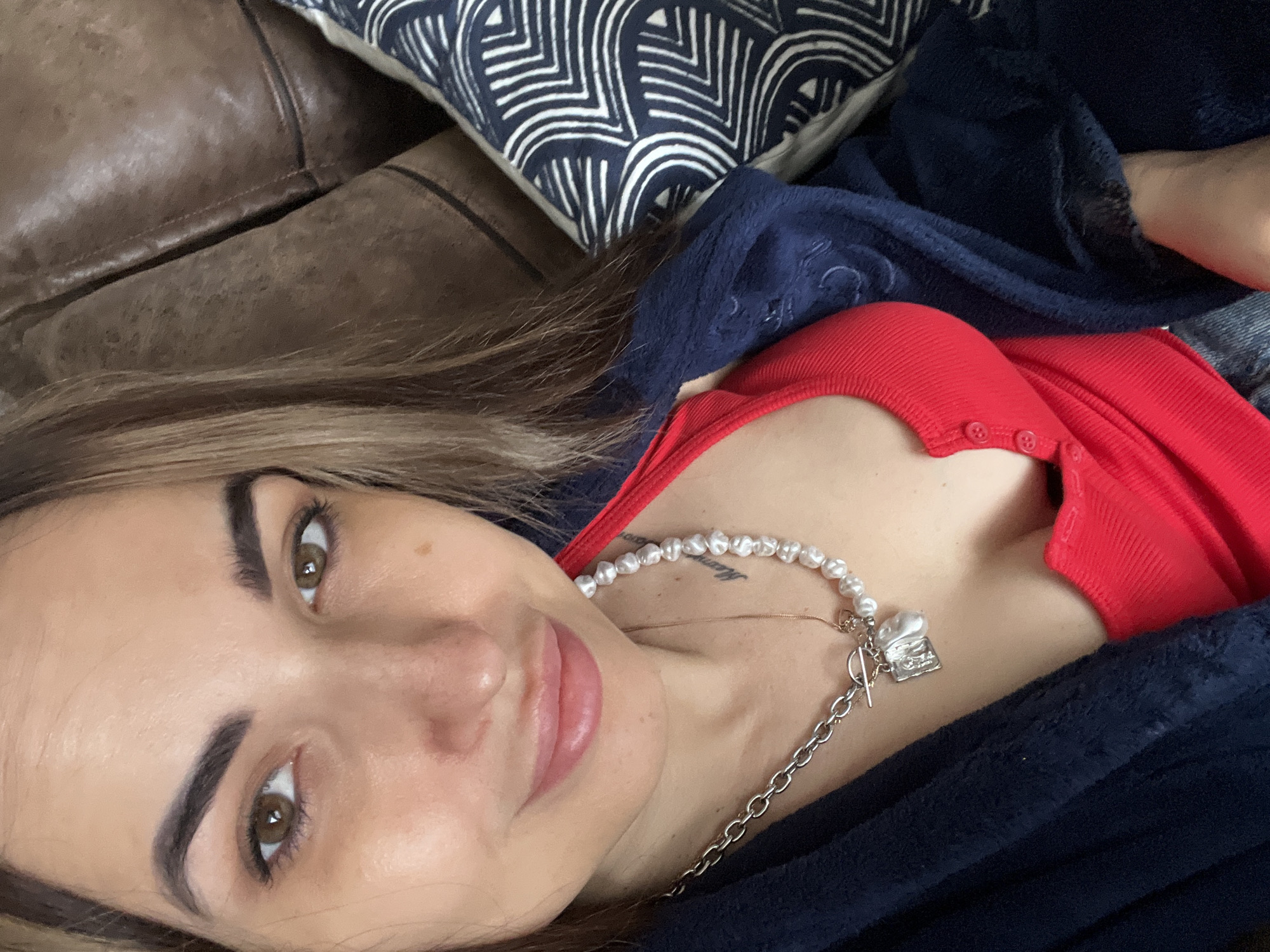 A close-up shot captures a person, likely a woman, lying down and looking upwards towards the camera. Her head is tilted, with light brown hair cascading around her. She has brown eyes and dark, well-defined eyebrows, and her lips are slightly parted, indicating a subtle, relaxed expression.
She is wearing a bright red, ribbed top with several small buttons down the front. A dark blue, soft-looking blanket or robe is draped over her, partially covering her torso and arms. A glimpse of blue jeans is visible on her right side. Around her neck, she wears a distinctive necklace featuring irregularly shaped white pearls on one side, transitioning into a silver chain with a toggle clasp on the other. A small, silver charm, possibly shaped like a miniature book or box, hangs from the chain, along with another thin gold chain layered beneath. A faint, cursive tattoo is partially visible on her upper chest, near her collarbone, but the text is illegible.
The setting appears to be indoors, suggesting a relaxed environment. Behind her, a rich brown leather surface, likely a couch or chair, is visible. Adjacent to the leather, a decorative cushion with a bold blue and white geometric pattern adds a touch of design to the background. The lighting is soft and diffused, characteristic of indoor ambient light, and there are no specific cues for the time of day. The overall impression is one of calm and comfort.