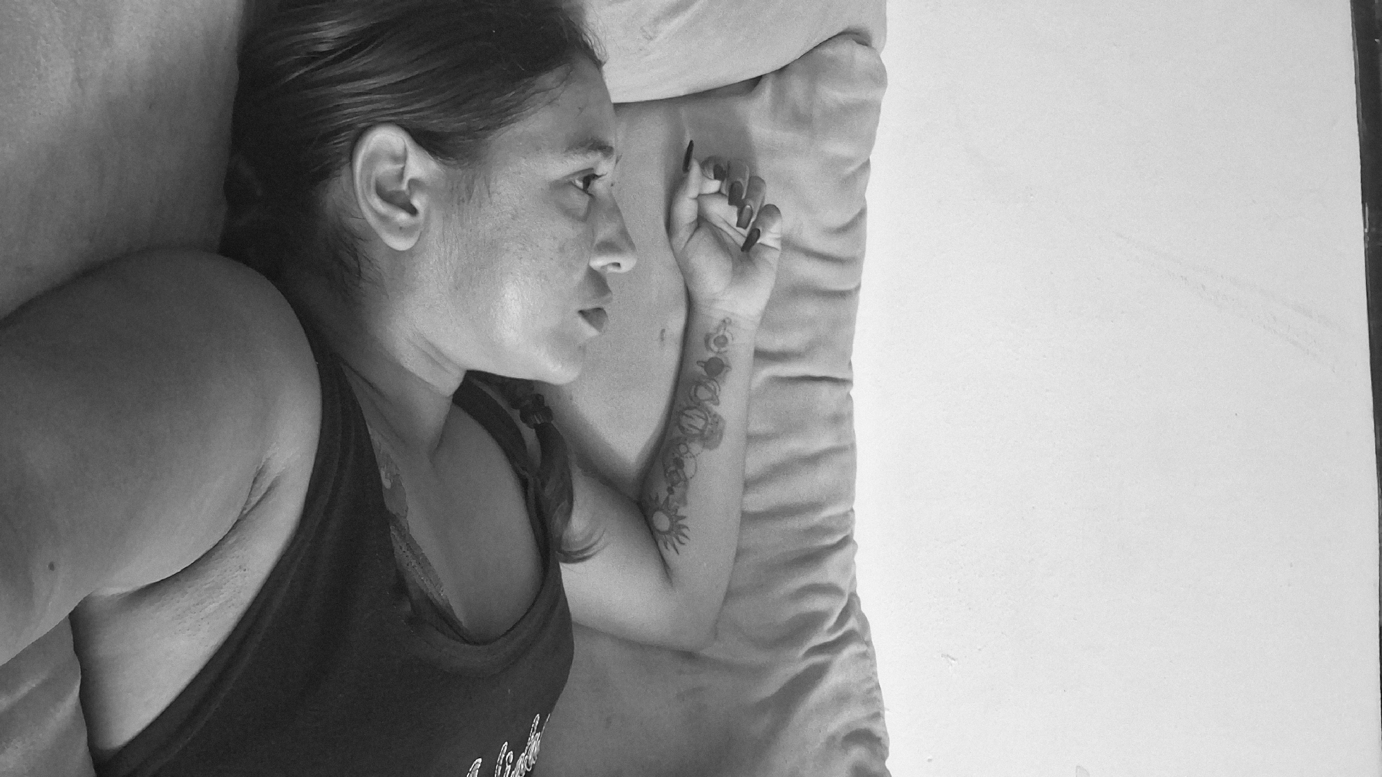 A black and white, vertical, medium-shot photograph shows a woman lying on her back on a light-colored sofa. She is looking upwards, her head tilted back. Her left arm is extended, with her hand resting near her head. The arm features a tattoo that appears to be a series of circular shapes and celestial bodies. She has long, dark fingernails. She is wearing a dark, sleeveless top. The background consists of a plain, light-colored wall and the textured surface of the sofa. The scene is indoors, likely in a residential setting within the city of Vallecito, Venezuela.