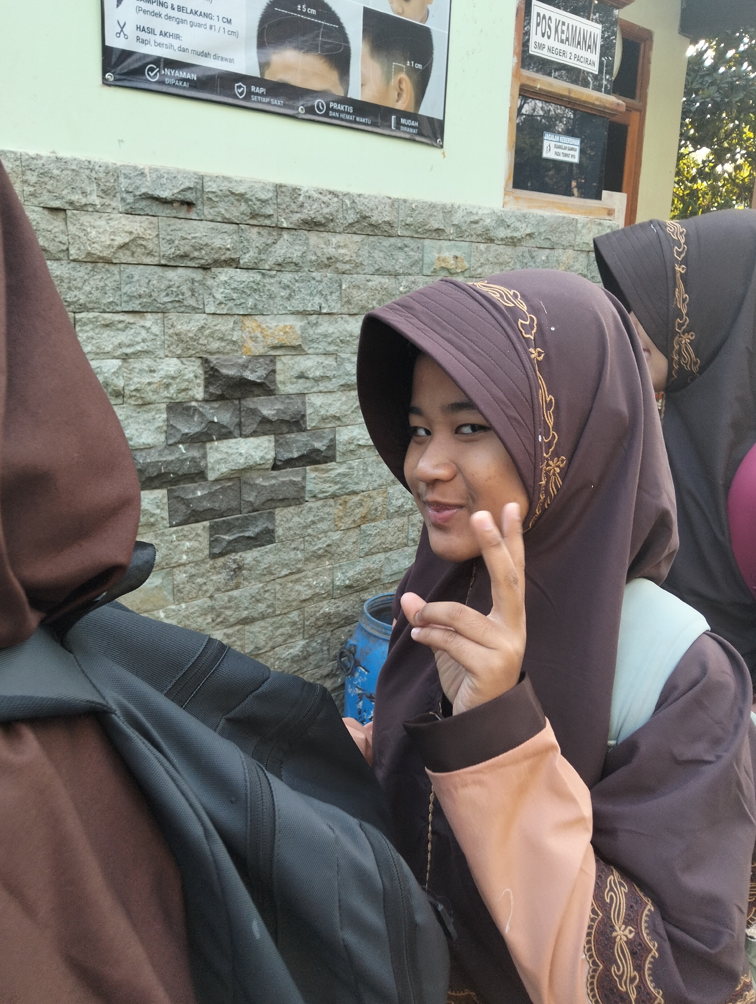 In the city of Paciran, Indonesia, two young women in brown hijabs are pictured outdoors. The woman in the foreground, facing the camera, is making a peace sign with her right hand. Her hijab is adorned with gold embroidery. She is wearing a peach-colored sleeve on her right arm. A black backpack is visible on her left side. Behind her, another young woman is partially visible, also wearing a brown hijab. The background consists of a stone-patterned wall and a light green wall with signage, including a sign that reads "POS KEAMANAN SMP NEGERI 2 PACIRAN." A blue barrel is visible to the right of the foreground woman. The scene appears to be in a school or public area.