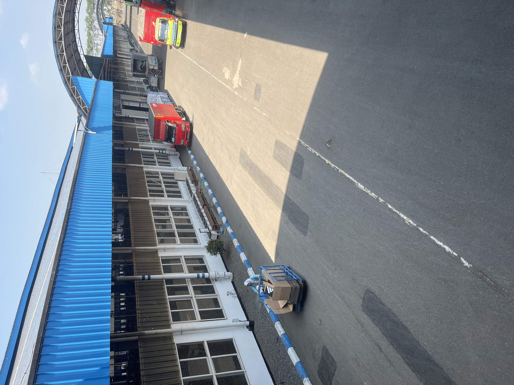 The image depicts an outdoor scene at a facility in the city of Pelang, Indonesia, likely a distribution center or factory yard. A blue-roofed building with white walls and numerous windows stretches across the left side of the frame. A person wearing a light blue headscarf and white clothing is seen pushing a blue cart filled with cardboard boxes along an asphalt pathway. The pathway is bordered by a curb with blue and white markings and some gravel. Two trucks are parked on the right side of the pathway: a red truck and a yellow truck. The red truck has a white logo on its side. The yellow truck has the word "KOPANA" visible on its side. The sky is clear and blue, indicating a sunny day. Shadows from the building and possibly other structures are cast across the asphalt.