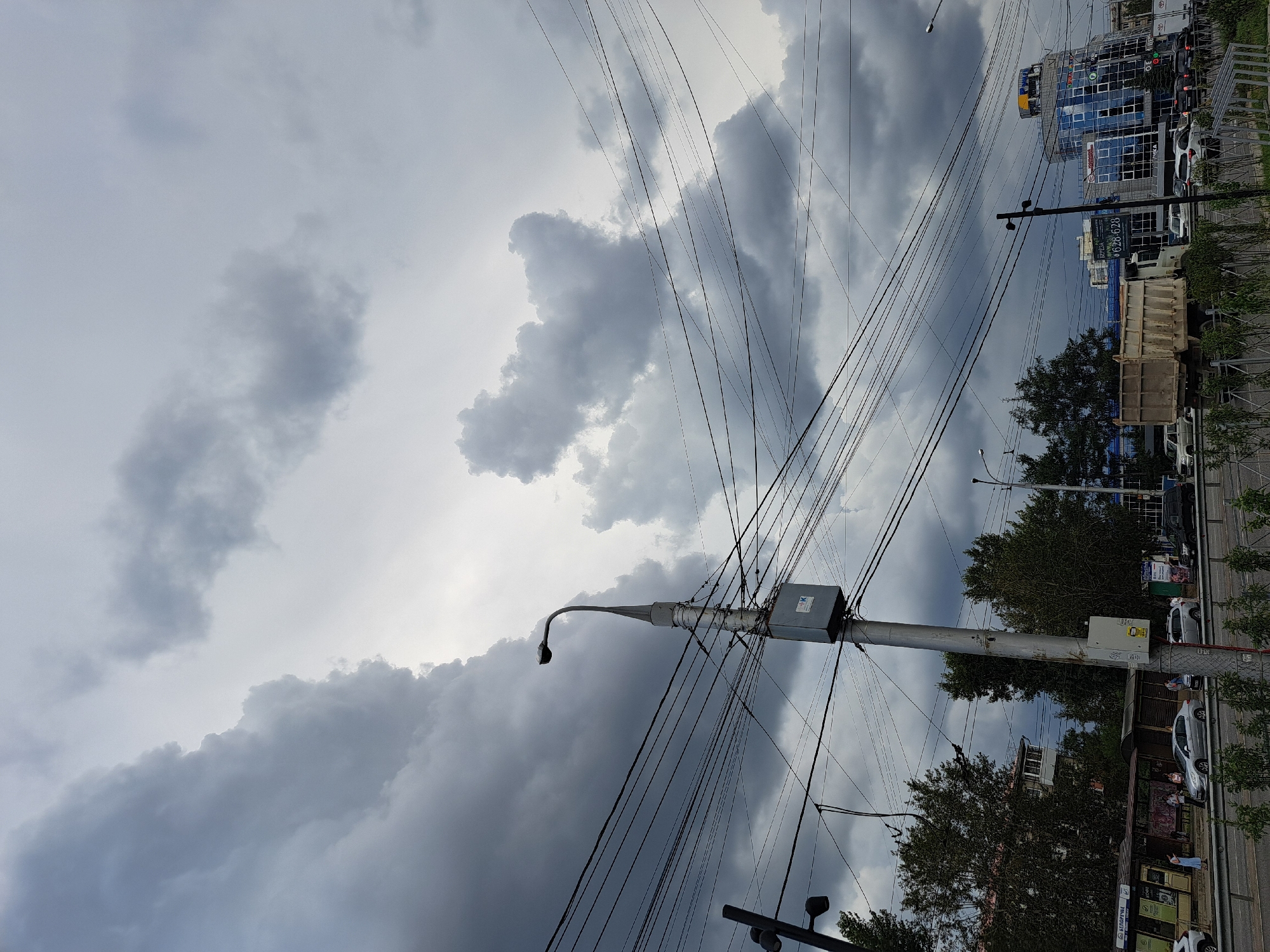 In Irkutsk, Russia, a street scene unfolds beneath a dramatic sky filled with dark, heavy clouds. The foreground is dominated by a utility pole adorned with numerous electrical wires, which crisscross the frame, creating a complex, almost tangled network. To the left, a smaller, darker pole with two lights is visible.
Further back, trees line the side of a road, their foliage dense and green. Several buildings are situated behind the trees, including a modern, blue-glass building with a yellow circular sign at its top. A light-colored dump truck is parked on the road, alongside other vehicles including cars and vans.
People are visible near a bus stop shelter on the left side of the image, some standing and others walking. The overall atmosphere is one of impending weather, with the turbulent clouds suggesting a storm. The scene appears to be captured from a roadside perspective, looking towards a busy urban area.