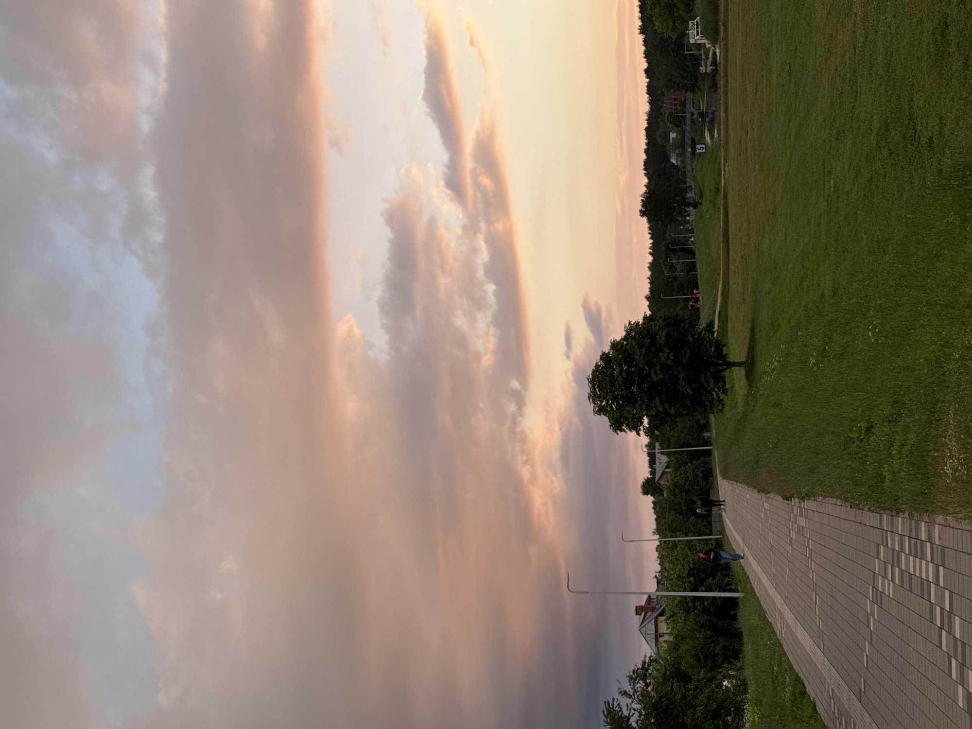 A paved walkway curves through a grassy park with a tree in the foreground and distant trees and buildings. The sky is filled with soft, colorful clouds in hues of pink, orange, and blue. Two people walk on the path in the middle ground, and several streetlights line the walkway. A small body of water with a bridge and some boats is visible in the distance. The scene appears to be set in a park in Carnikava, Latvia, during a sunset.