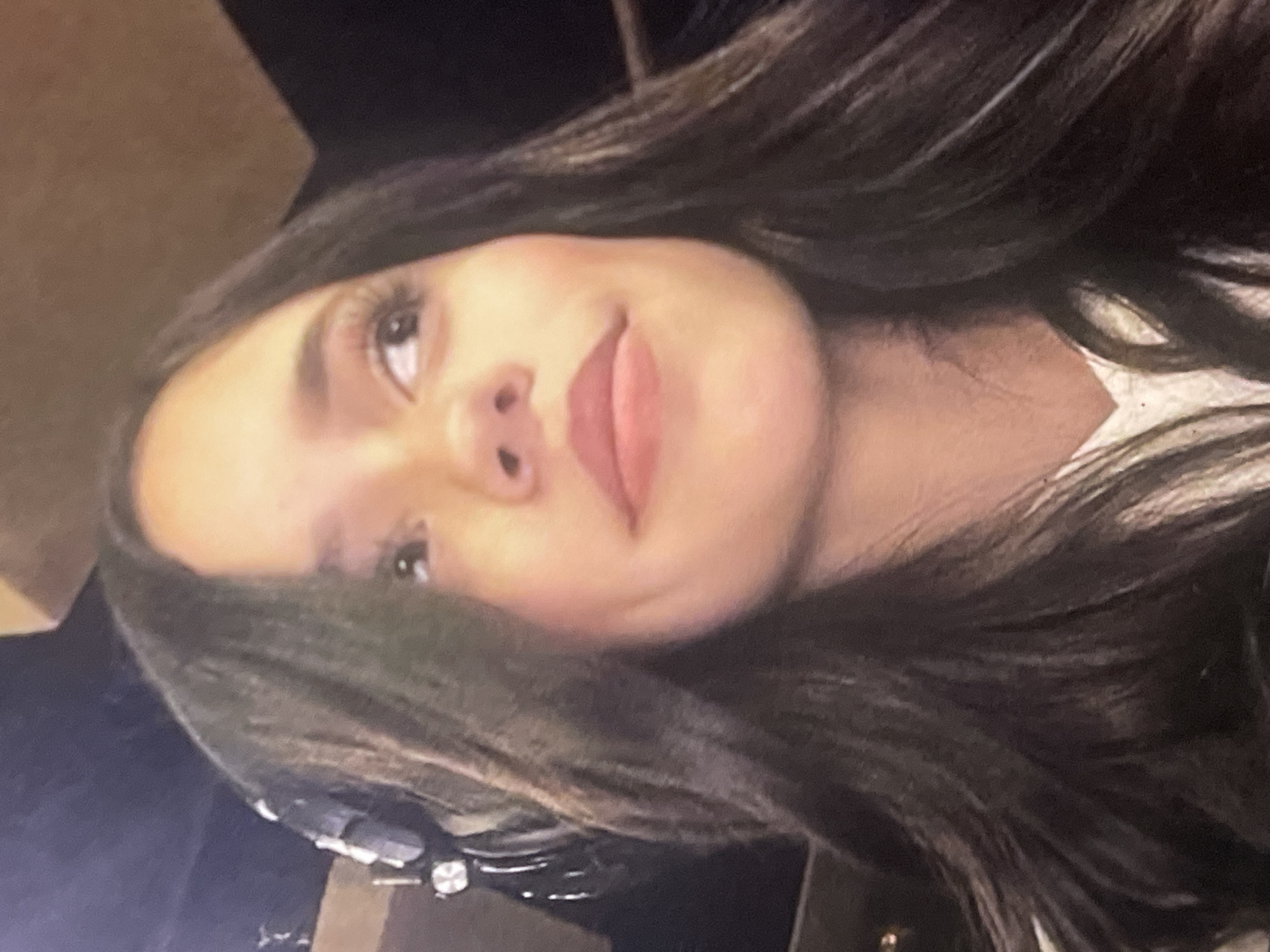 This is a close-up portrait of a young woman, with the image rotated 90 degrees clockwise from a standard upright orientation. She has long, dark hair that frames her face and flows downwards. Her dark eyes are looking slightly upwards and to her left (relative to the rotated image). She has prominent eyelashes, and her eyebrows are well-defined. Her lips are full and slightly parted, revealing a subtle, gentle smile or smirk. Her complexion appears smooth and clear.
She is wearing a light-colored garment, possibly white, visible on her right side (her right shoulder/chest area in the rotated image). On the opposite side, beneath her chin, there is a dark, indistinct fabric, which might be part of another piece of clothing or an accessory like a headphone band. In the bottom-left corner of the rotated image, a small, shiny, metallic object, possibly jewelry or a watch, is partially visible.
The background is dark and out of focus, suggesting an indoor setting or an environment with low lighting. Hints of dark, possibly upholstered or wooden surfaces can be discerned in the blurred background. The woman's expression is calm and relaxed. There is no visible text in the image.