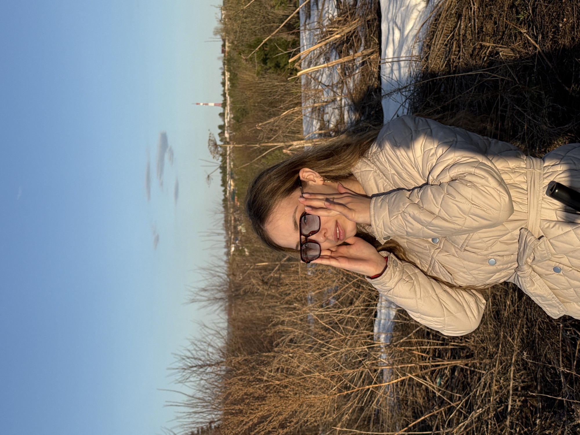 A woman lies on a field of dry grass and snow in Chaykovsky, Russia. The woman is wearing a beige quilted jacket and eyeglasses. She is holding her eyeglasses up with both hands. She is in the lower right of the frame. The background is a field of dry, brown grass and snow. In the distance, there is a cityscape with a tall, thin red and white chimney. The sky is a clear blue.